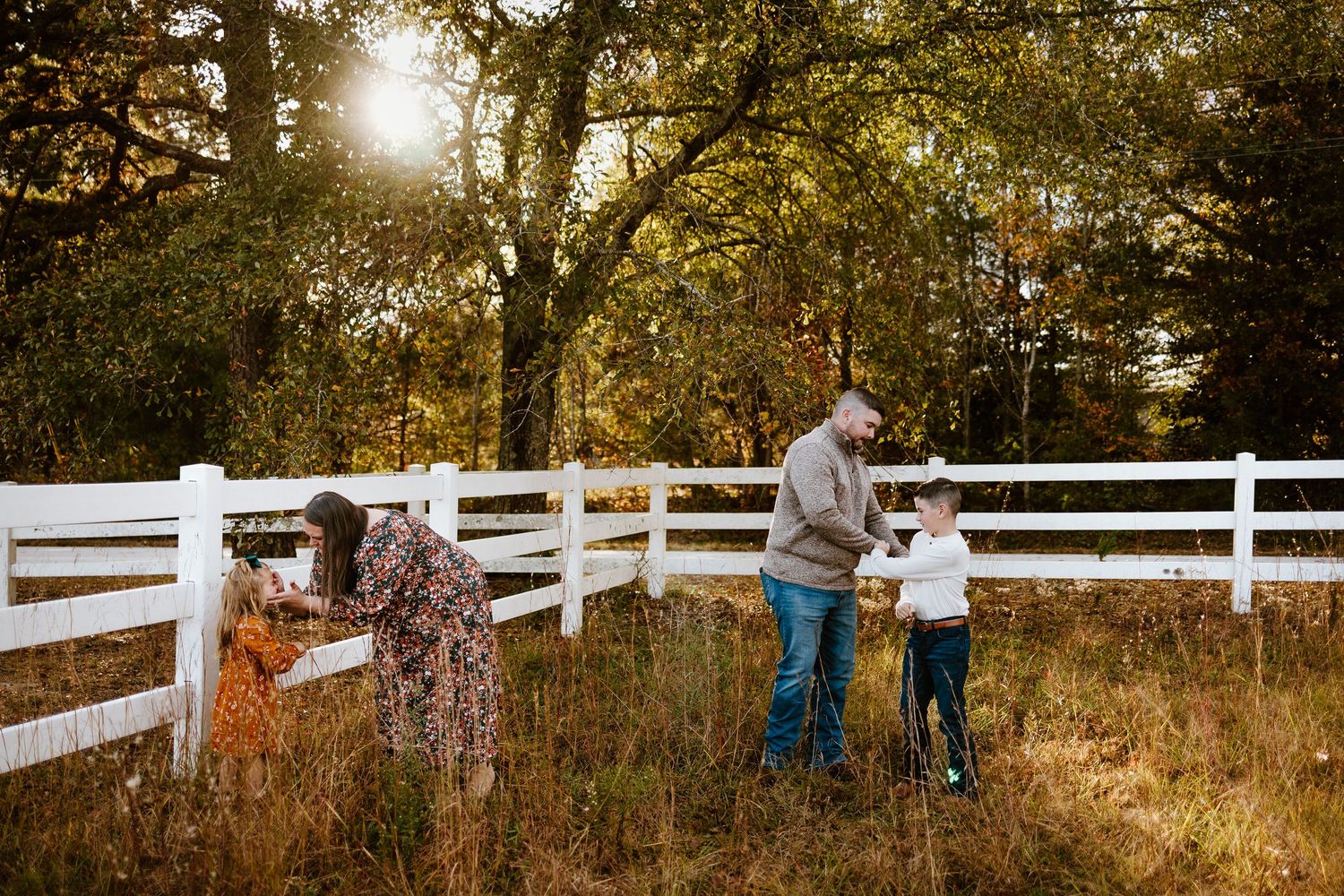 Family exploring a white fence in a golden autumn field surrounded by lush green trees at sunset.