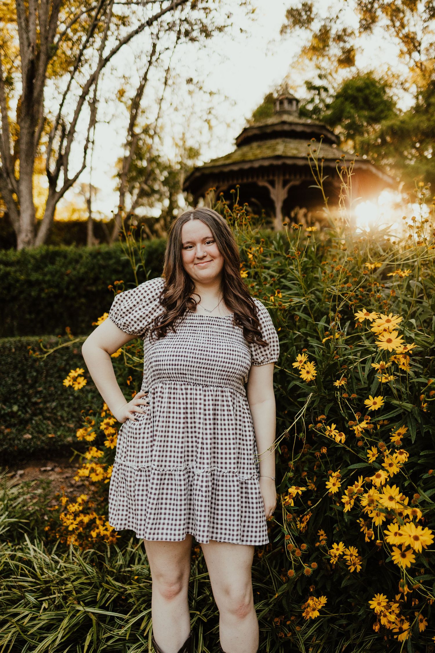 Smiling young woman in a patterned dress posing near yellow flowers with a gazebo at golden hour.