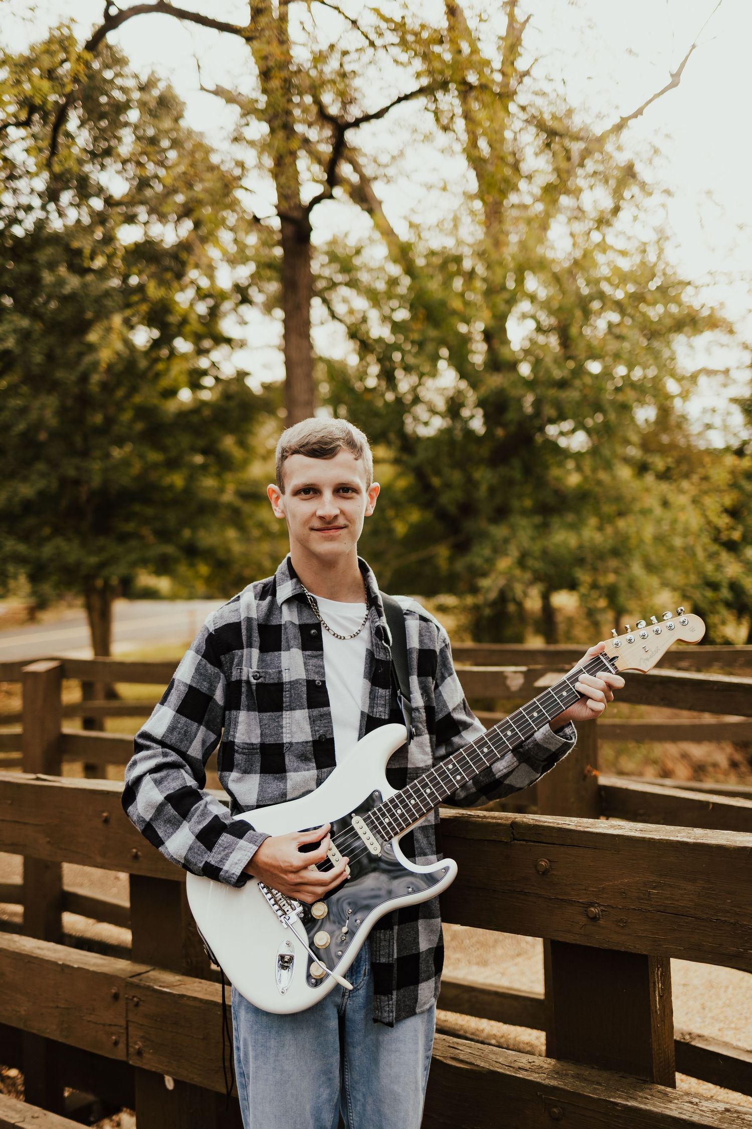 Young man in plaid shirt holding white electric guitar outdoors near wooden fence in fall setting.