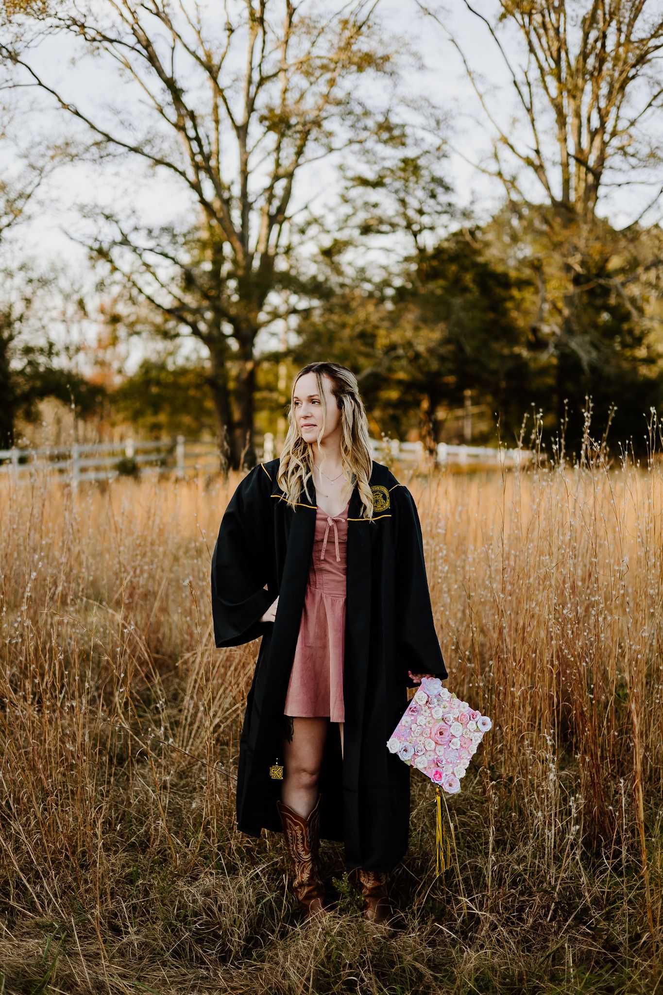 Graduate in black robe holding floral decorated cap standing in golden autumn field at sunset.