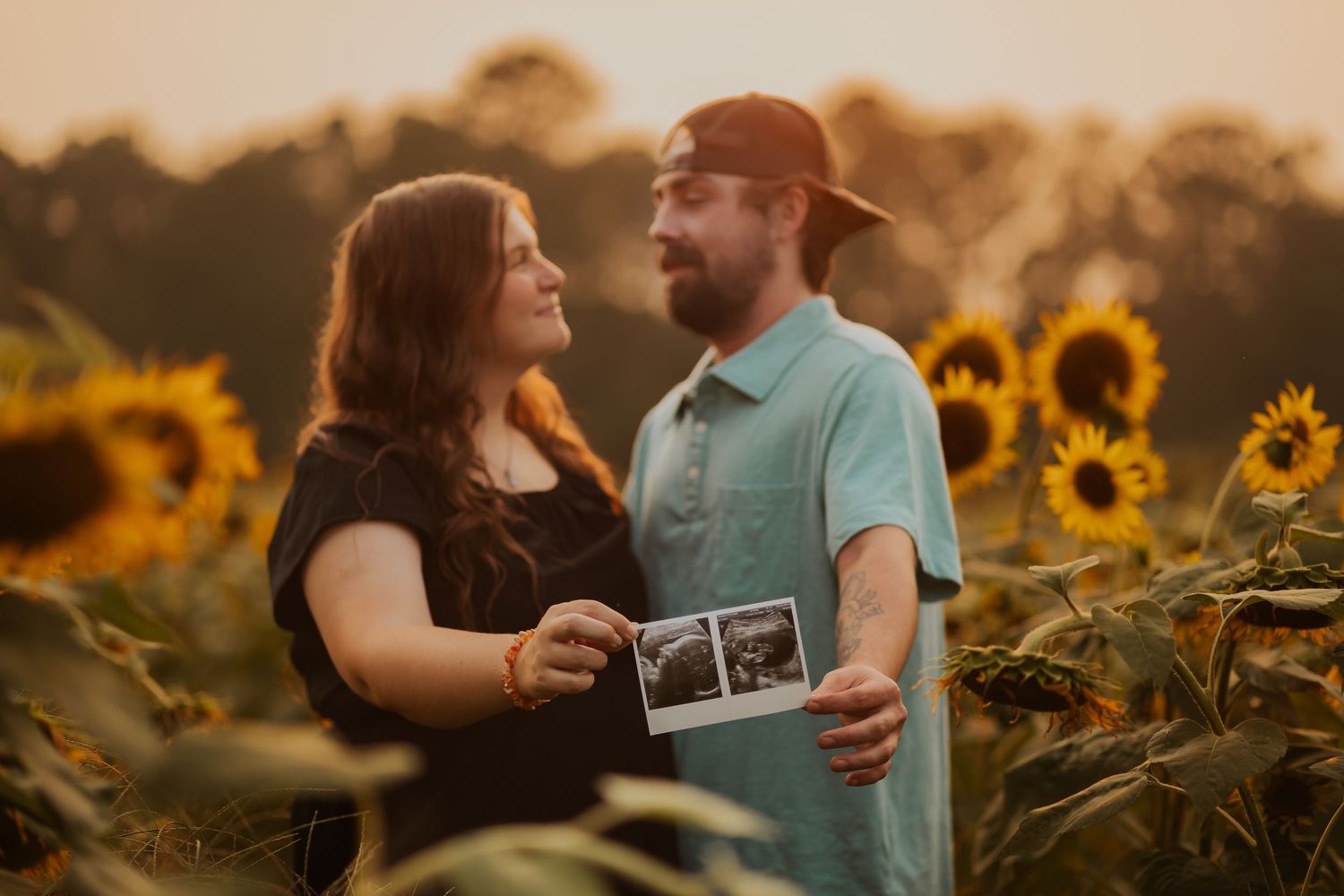 Couple shares a tender moment in a sunflower field while holding an ultrasound photo at golden hour.