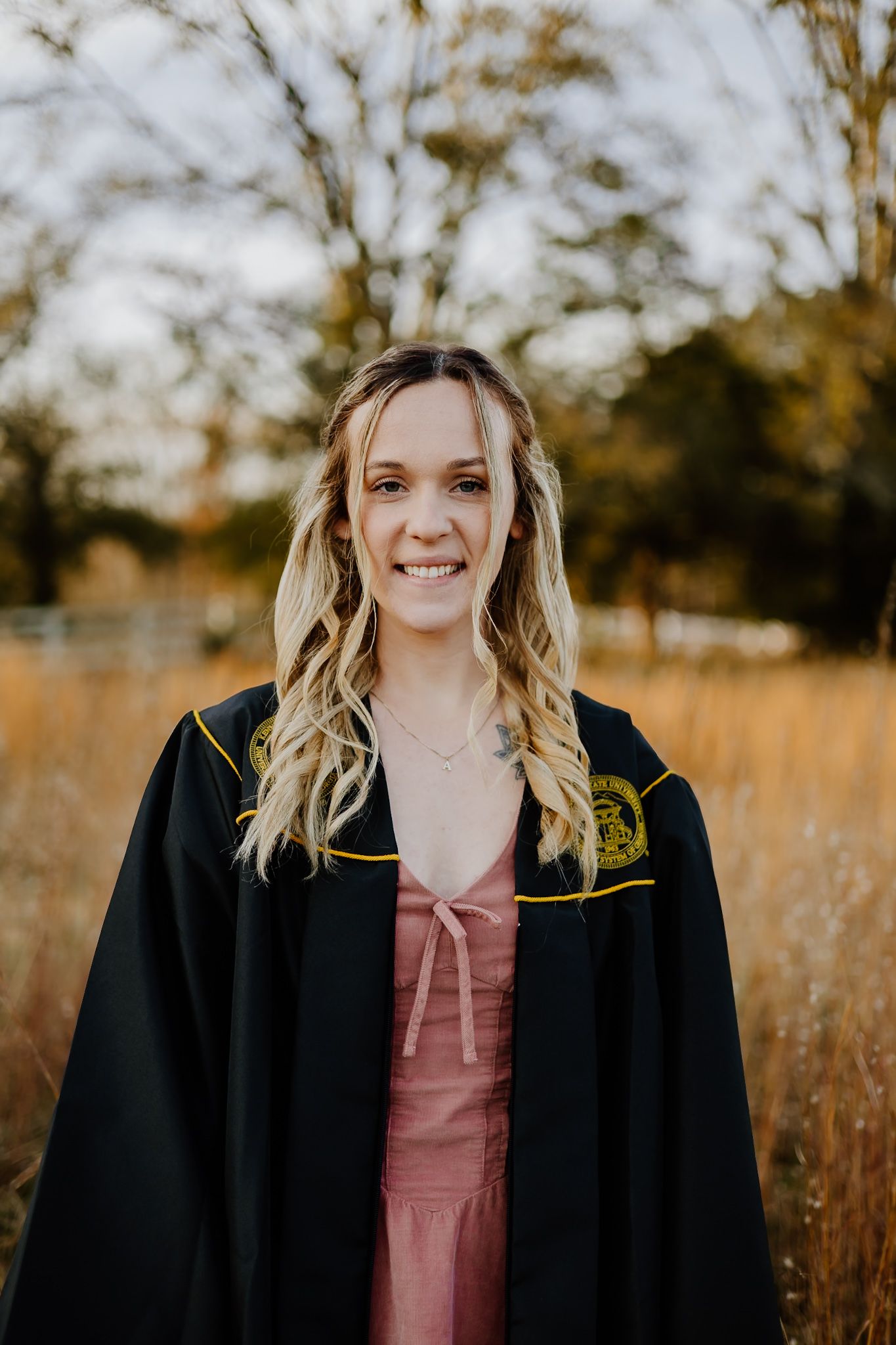 Smiling graduate in black academic gown with gold emblem standing in autumn outdoor setting.