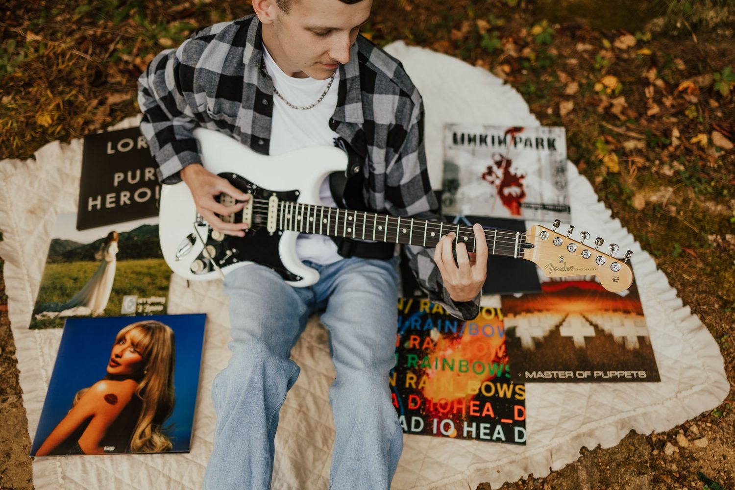 Teen boy playing white electric guitar surrounded by vinyl records including Linkin Park on white rug.