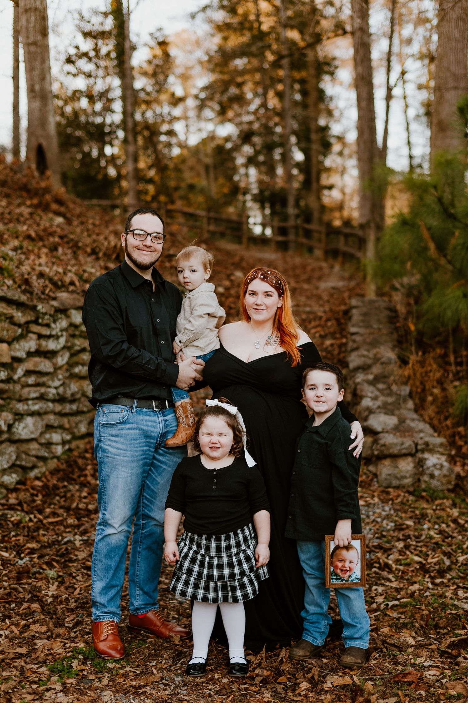 Family of five posing in autumn woods with stone wall and colorful fallen leaves.