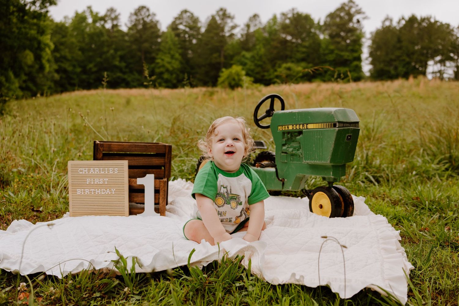 Baby's first birthday photo with John Deere tractor toy on white blanket in green meadow field.