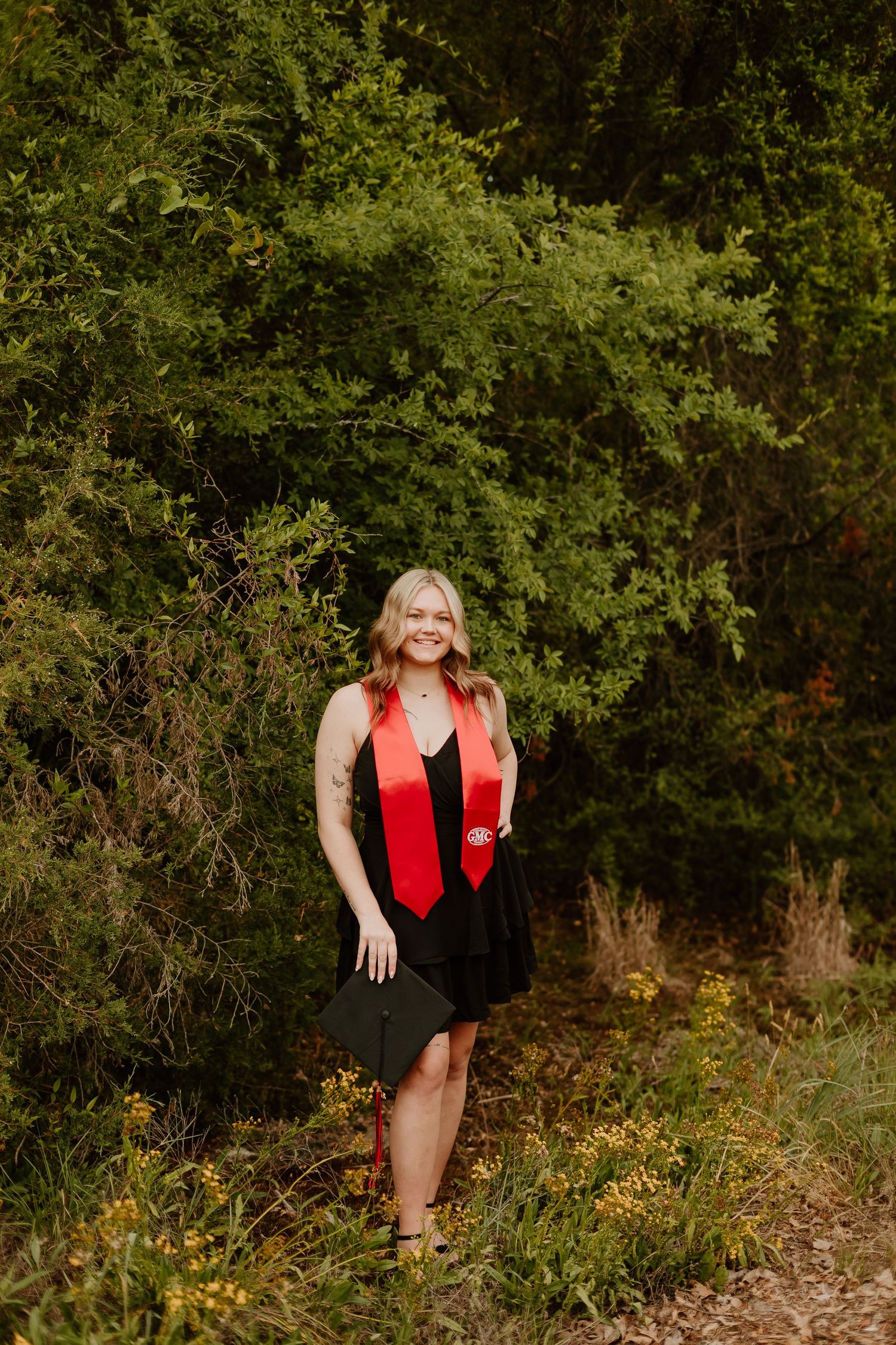 Smiling blonde graduate in black dress and red sash holds cap outdoors among lush green trees and wildflowers.