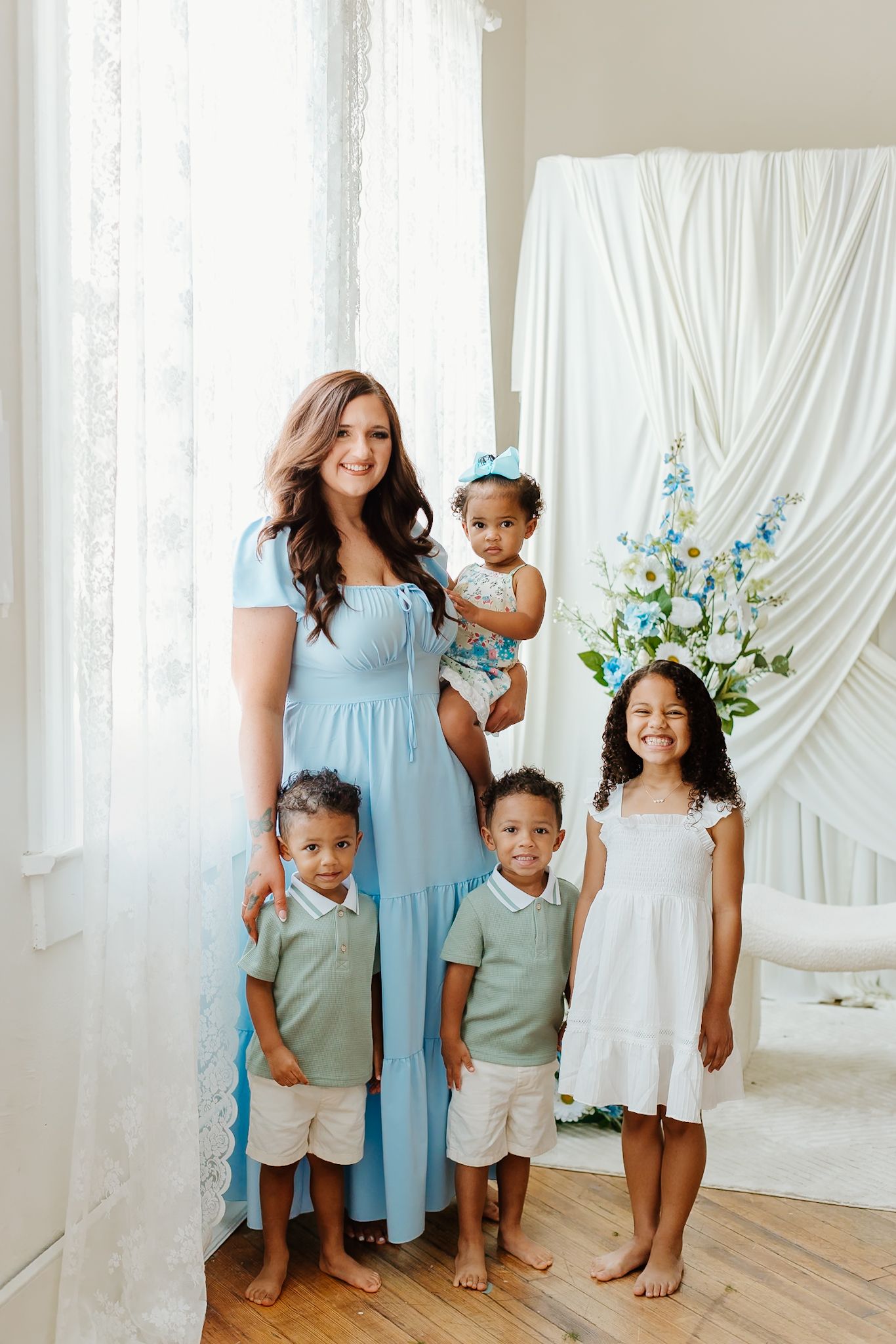 A woman in a light blue dress poses with four young children in a bright white studio setting with floral decor.