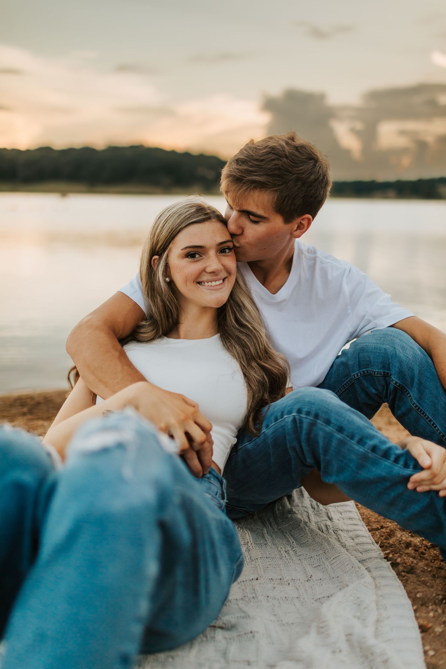 Couple sitting by a lake at sunset, man kissing woman on the cheek.