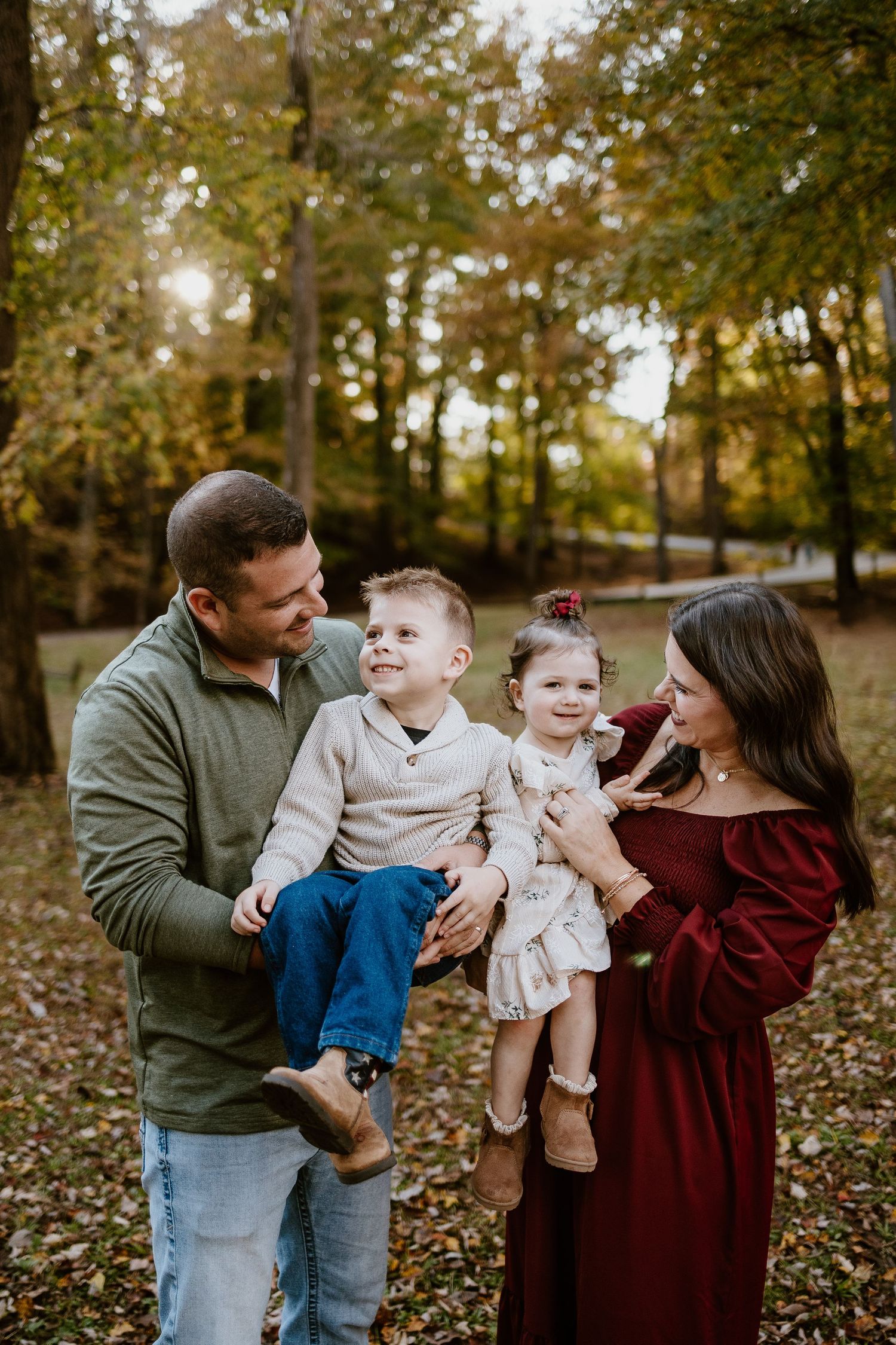 Smiling family of four with two toddlers enjoying autumn park portrait session among fall foliage.