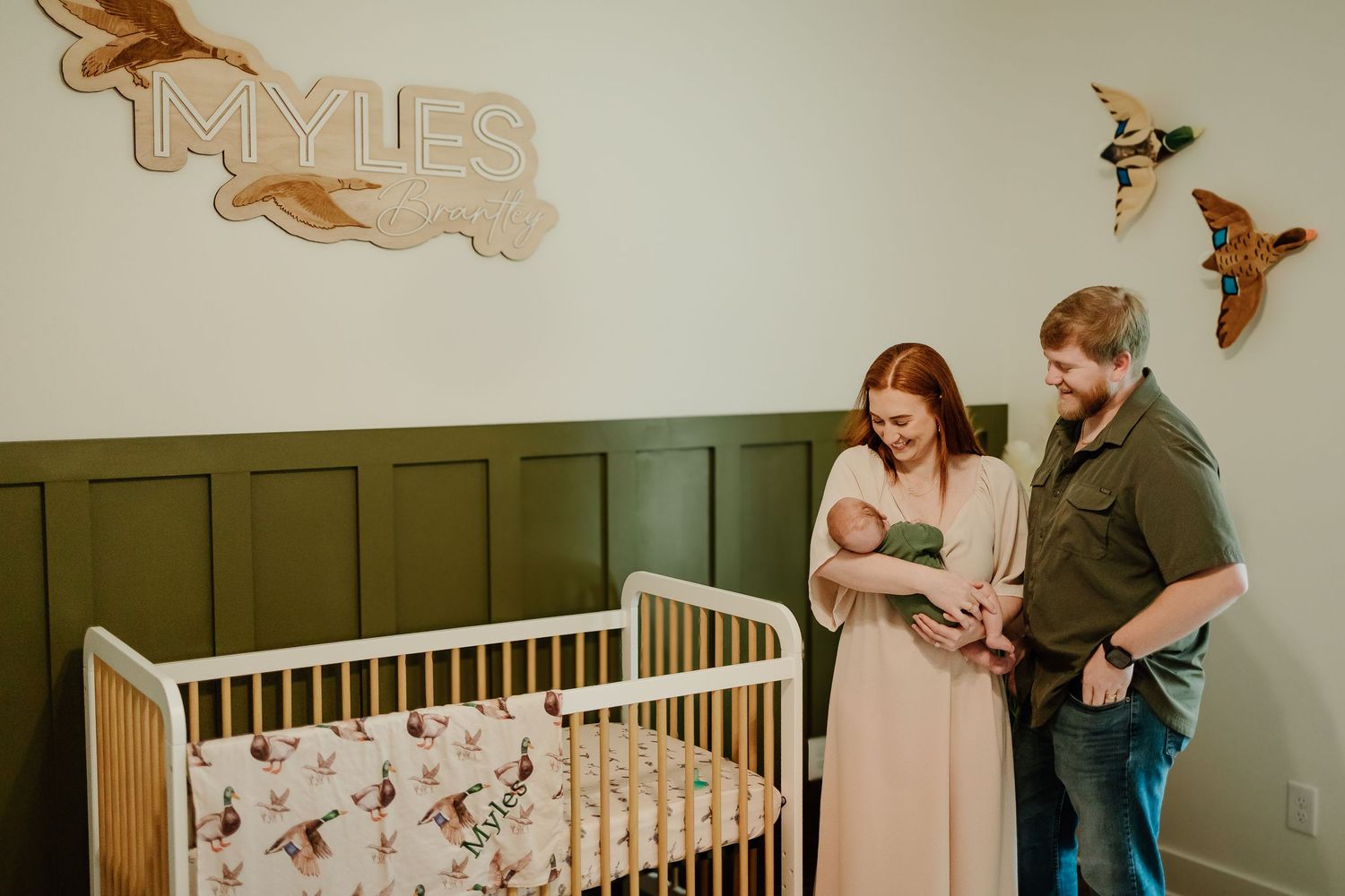 New parents admiring newborn baby in green nursery with custom name sign above crib.