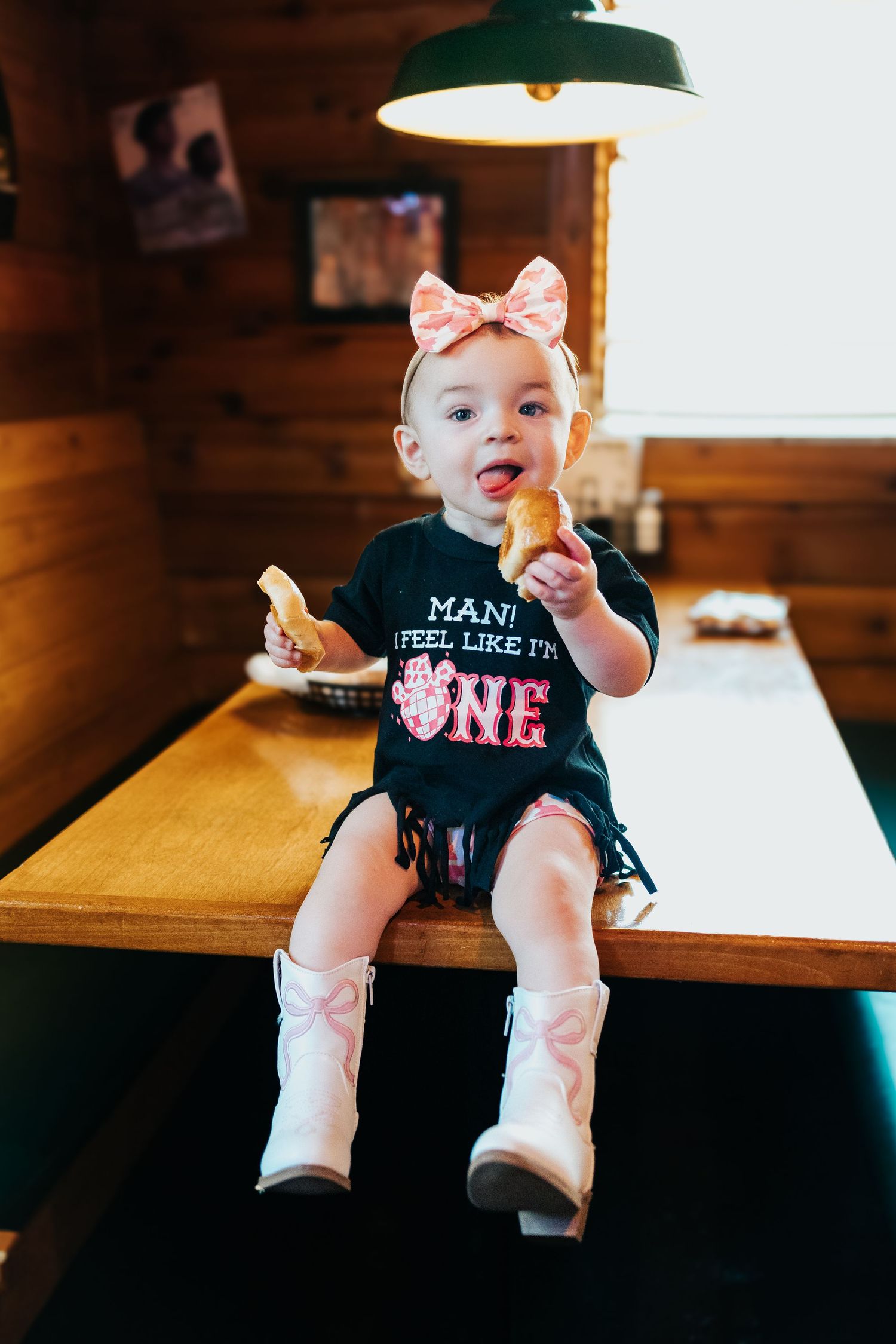 One-year-old girl in birthday shirt and white cowboy boots sits on table holding food with tongue out.