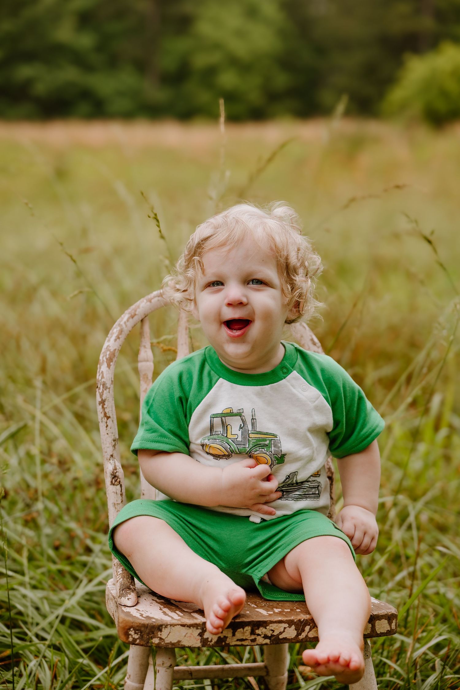 Laughing toddler boy in green outfit sitting on rustic chair in grassy field during birthday photoshoot.