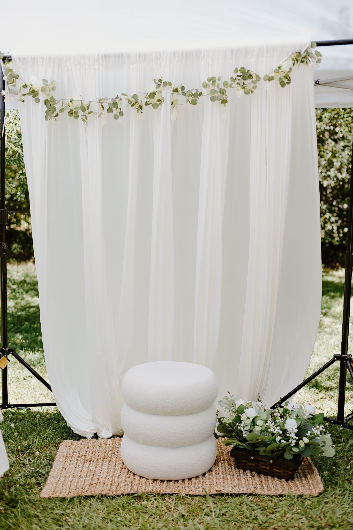 Elegant outdoor wedding backdrop with white draping, greenery garland, stacked ottoman, and floral arrangement on woven rug.