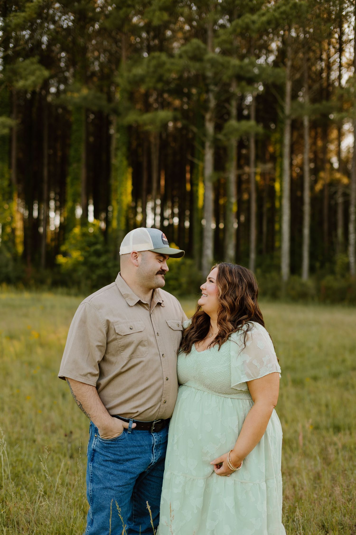 A couple shares a loving glance in a sunlit meadow, man in tan shirt and cap, woman in mint green dress.