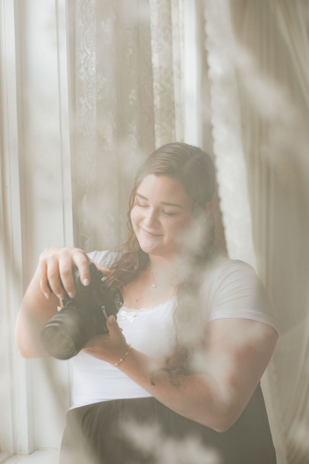 Woman in white top smiling while reviewing photos on her camera, standing by a sunlit window with sheer curtains.
