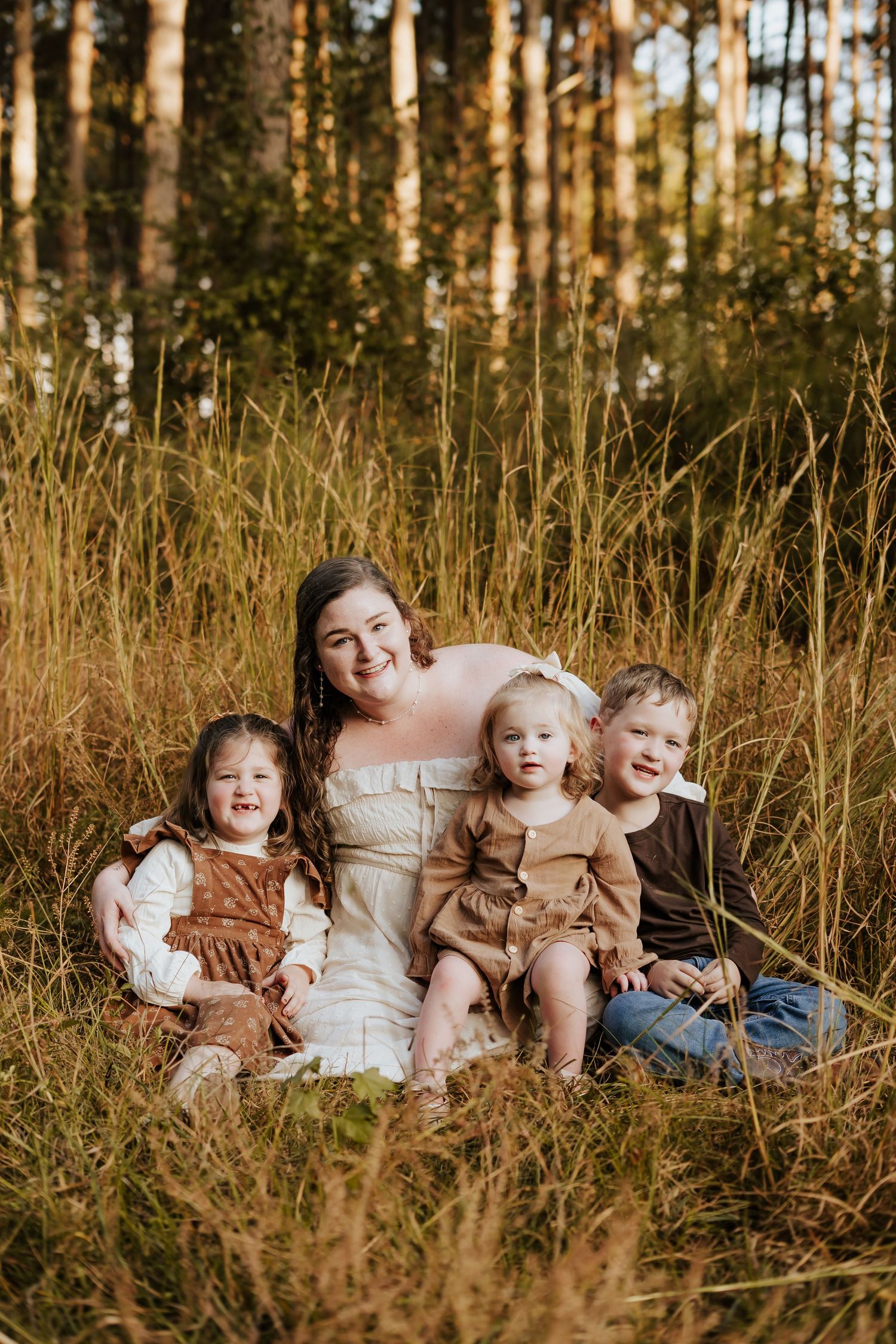 Mother in a white dress sitting in tall golden grass with her three young children in a pine forest.