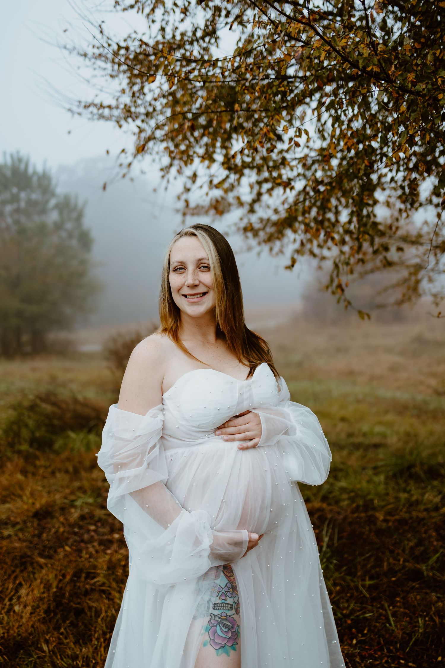 Smiling pregnant woman in sheer white maternity gown standing in misty autumn field.