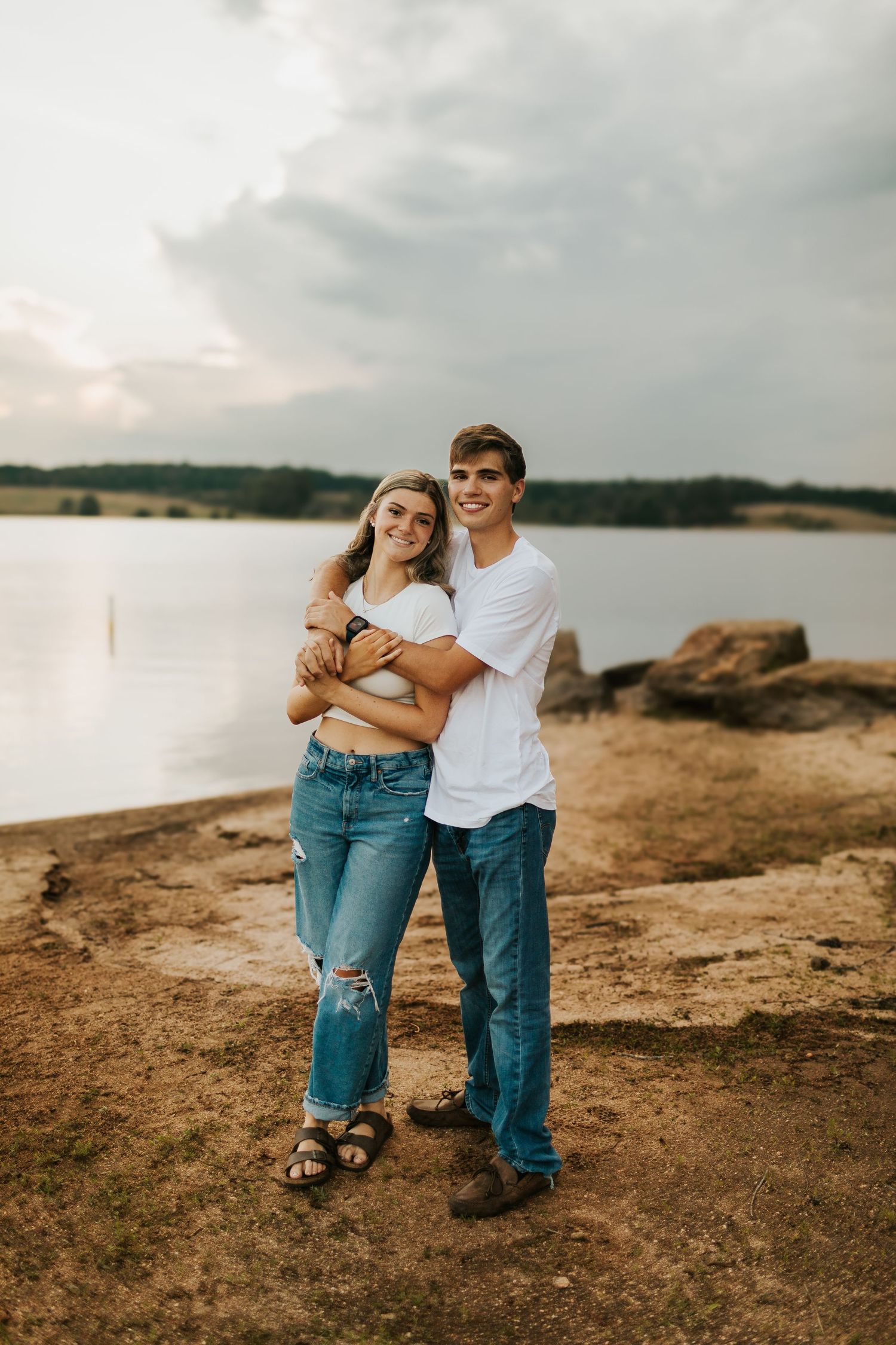 Young couple embracing and smiling together on a rocky lakeside at dusk.