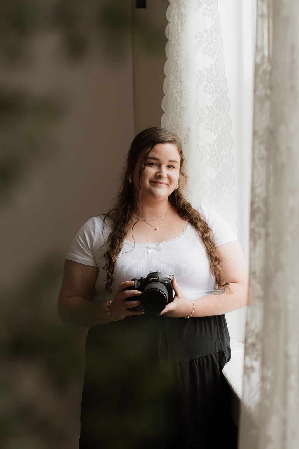 Young woman with long curly hair holding a DSLR camera, smiling by a lace-curtained window in white top and dark skirt.