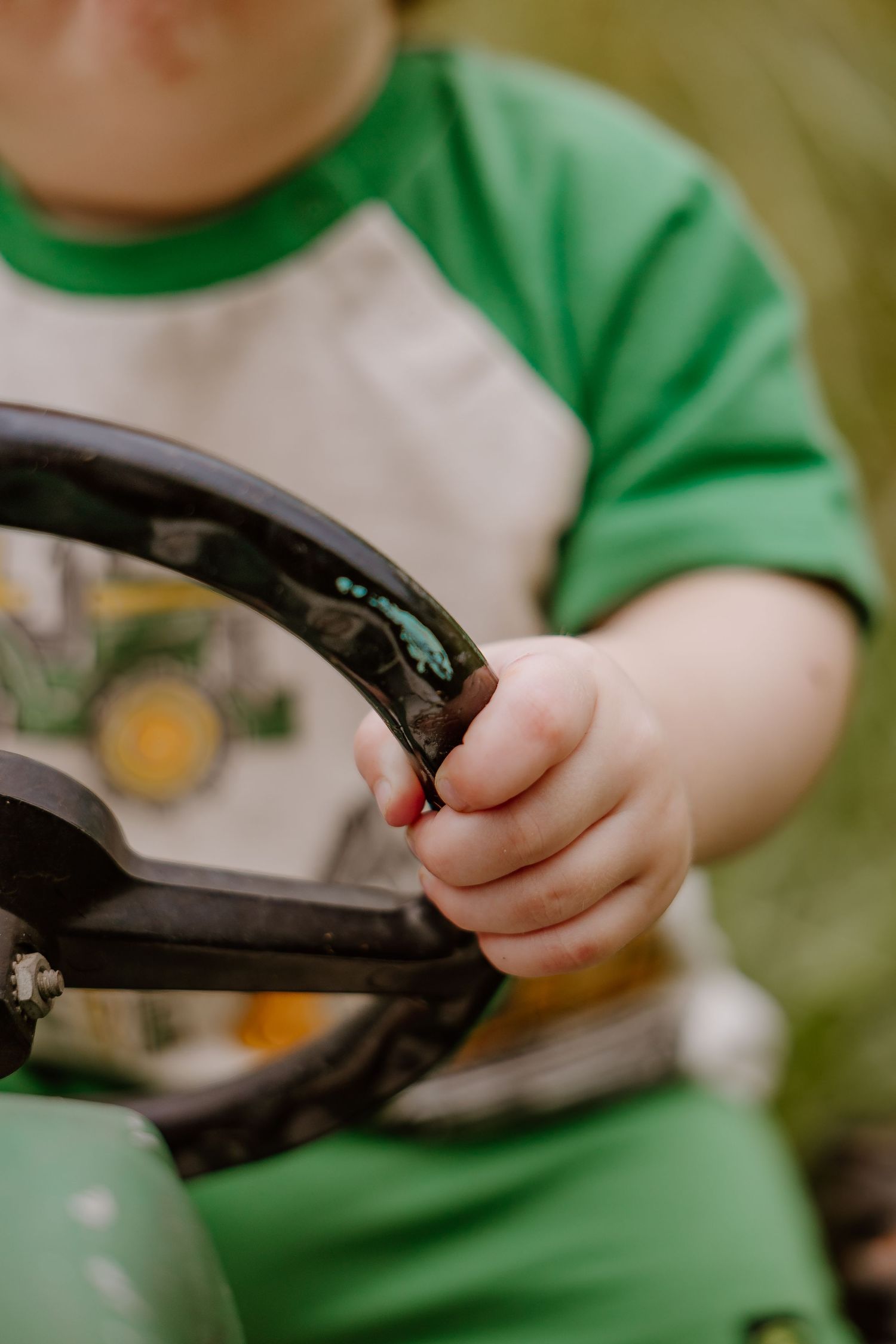 Young child in green shirt gripping a black steering wheel on a toy John Deere tractor outdoors.