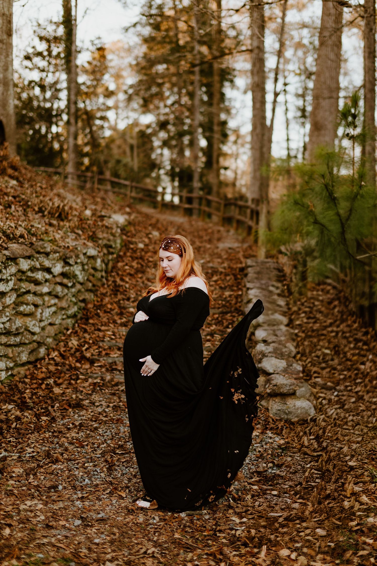 Pregnant woman in flowing black off-shoulder gown posing in autumn woodland setting.