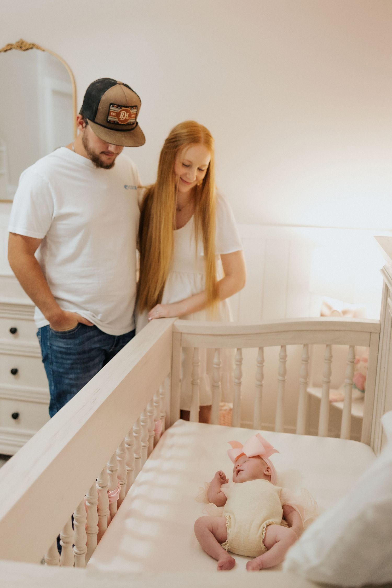 New parents lovingly gaze at their newborn baby lying in a white crib in a soft pink nursery.
