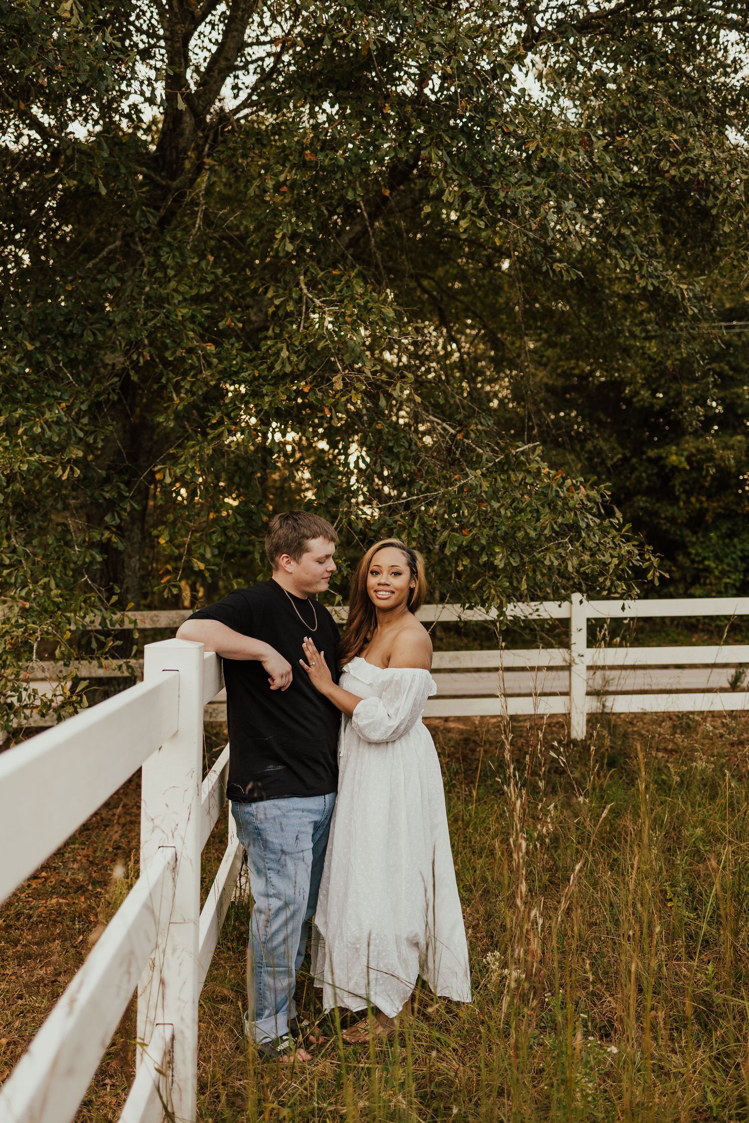 Couple standing together by a white fence in a lush green outdoor setting.
