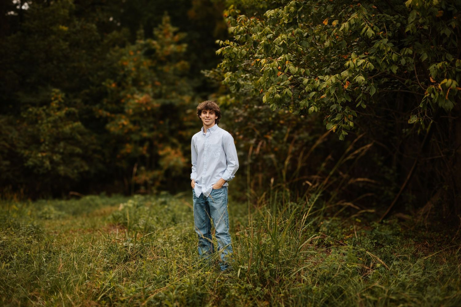 Senior boy in light blue shirt standing in lush green meadow surrounded by dense autumn trees.