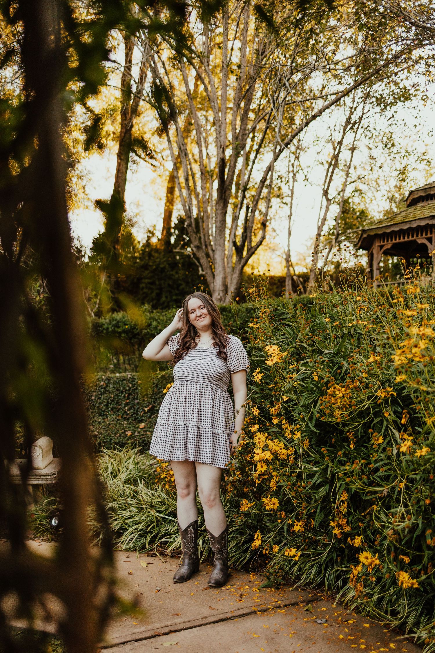 Young woman in gingham dress and cowboy boots posing beside yellow wildflowers in garden setting.