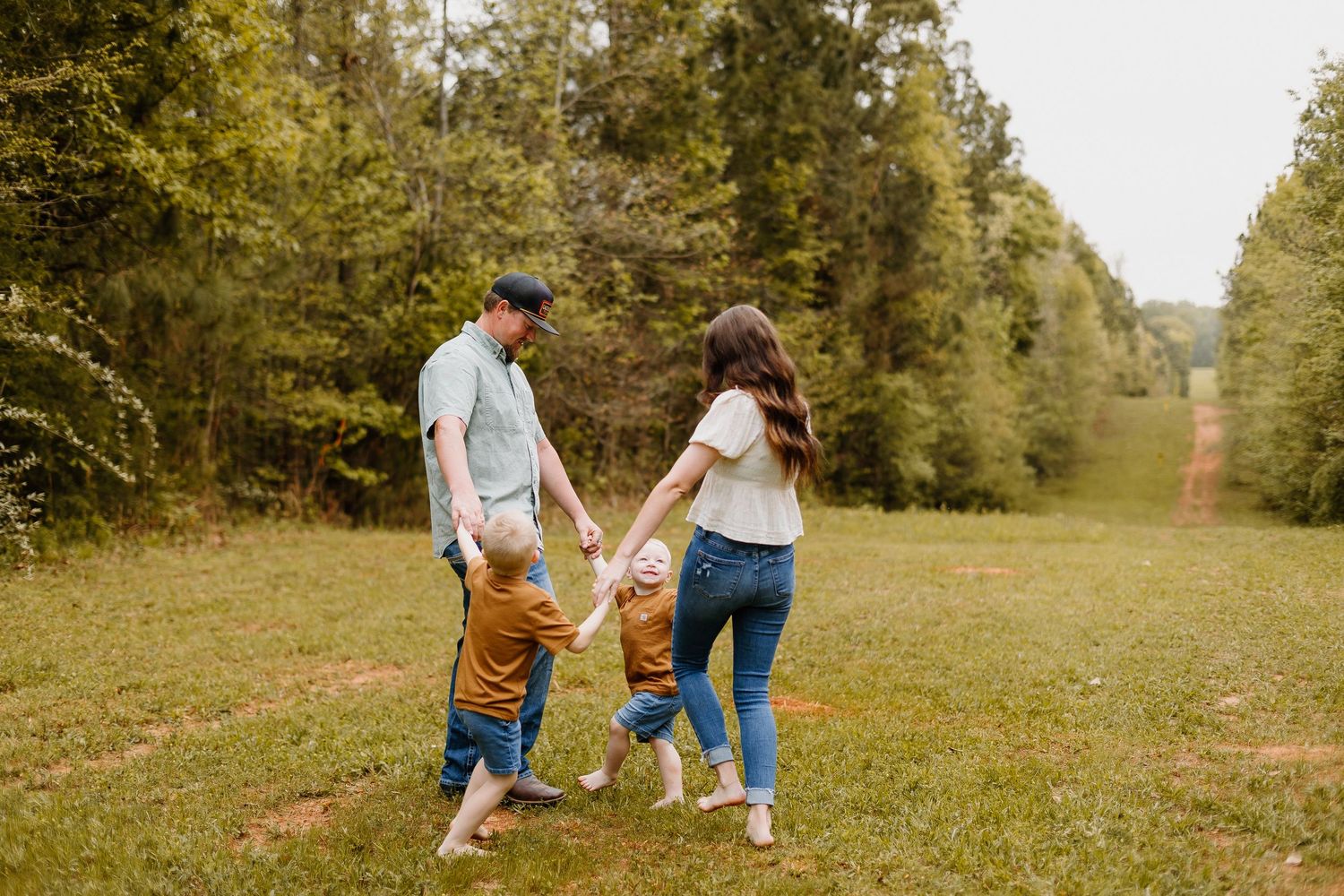 Family of four holding hands walking through a lush green meadow surrounded by trees.