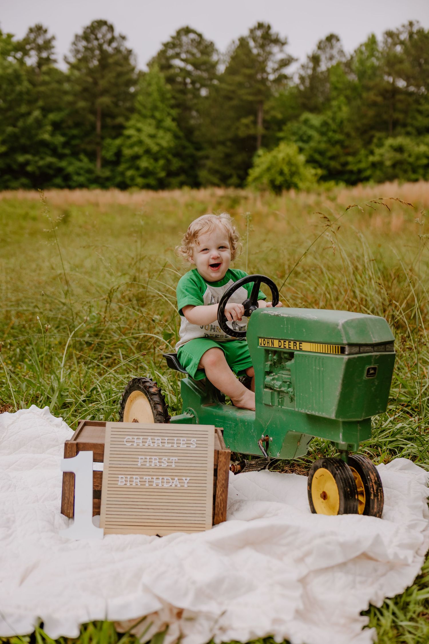 Happy toddler boy riding green John Deere pedal tractor at Charlie's first birthday photoshoot in field.