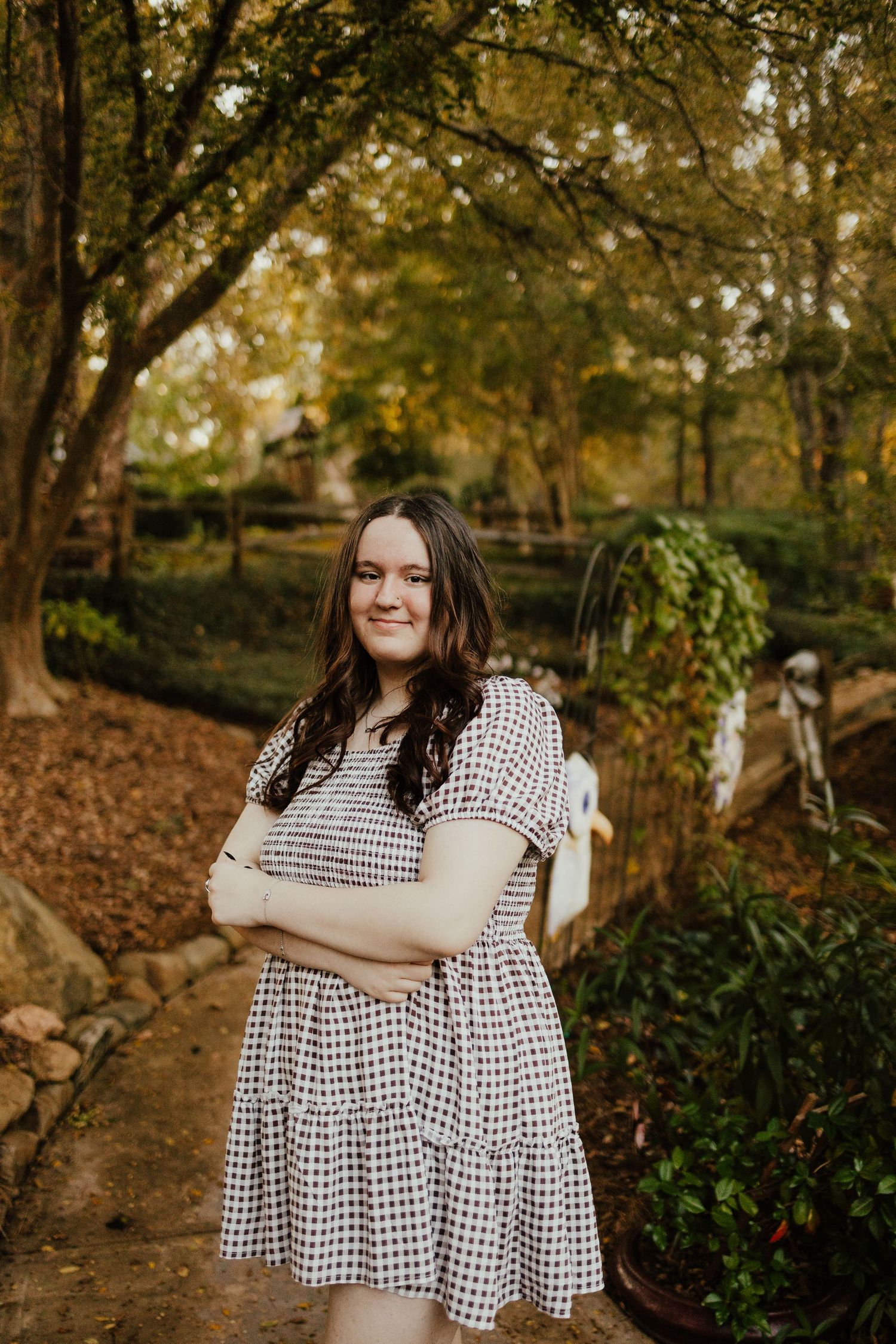 Young woman in gingham dress posing with arms crossed on garden path surrounded by autumn foliage.