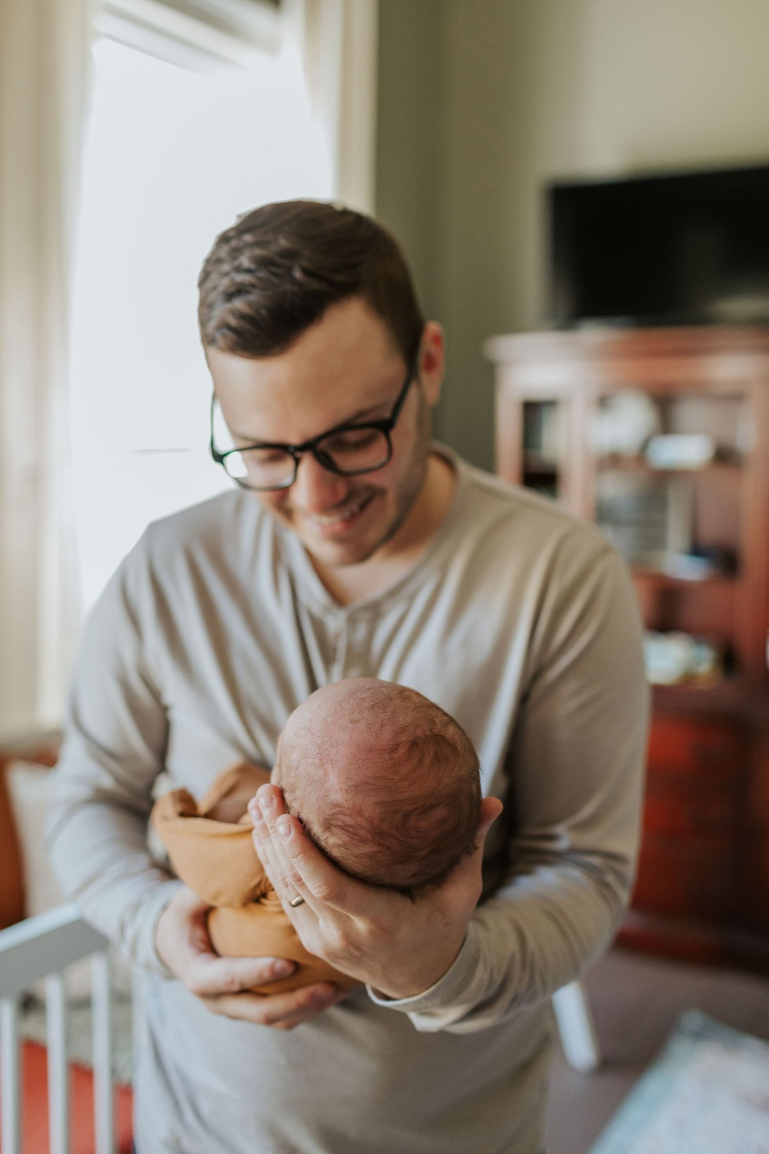 Smiling father with glasses tenderly holds newborn baby in a cozy home nursery setting.
