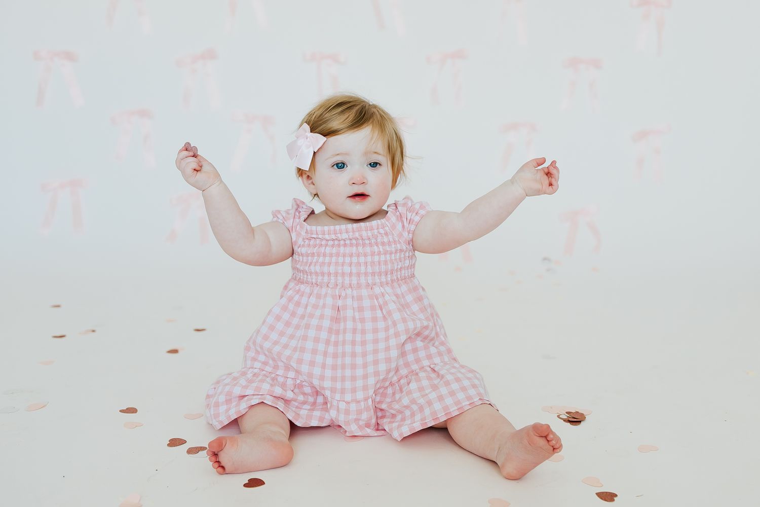 Adorable baby girl in pink gingham dress sitting on white floor with arms raised, wearing bow headband.