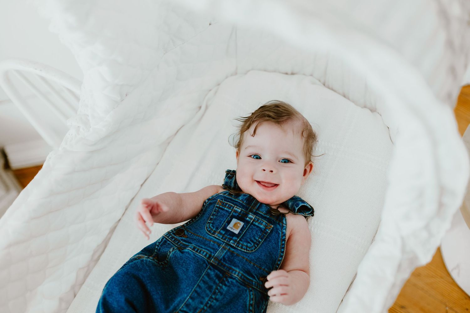 Smiling baby in denim Carhartt overalls lying in white bassinet during lifestyle newborn session.