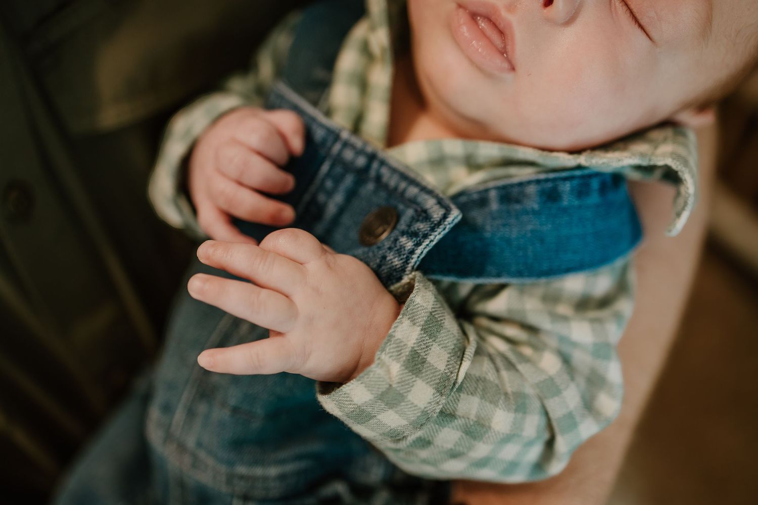 Close-up of newborn baby wearing a plaid shirt and denim overalls with tiny delicate hands.