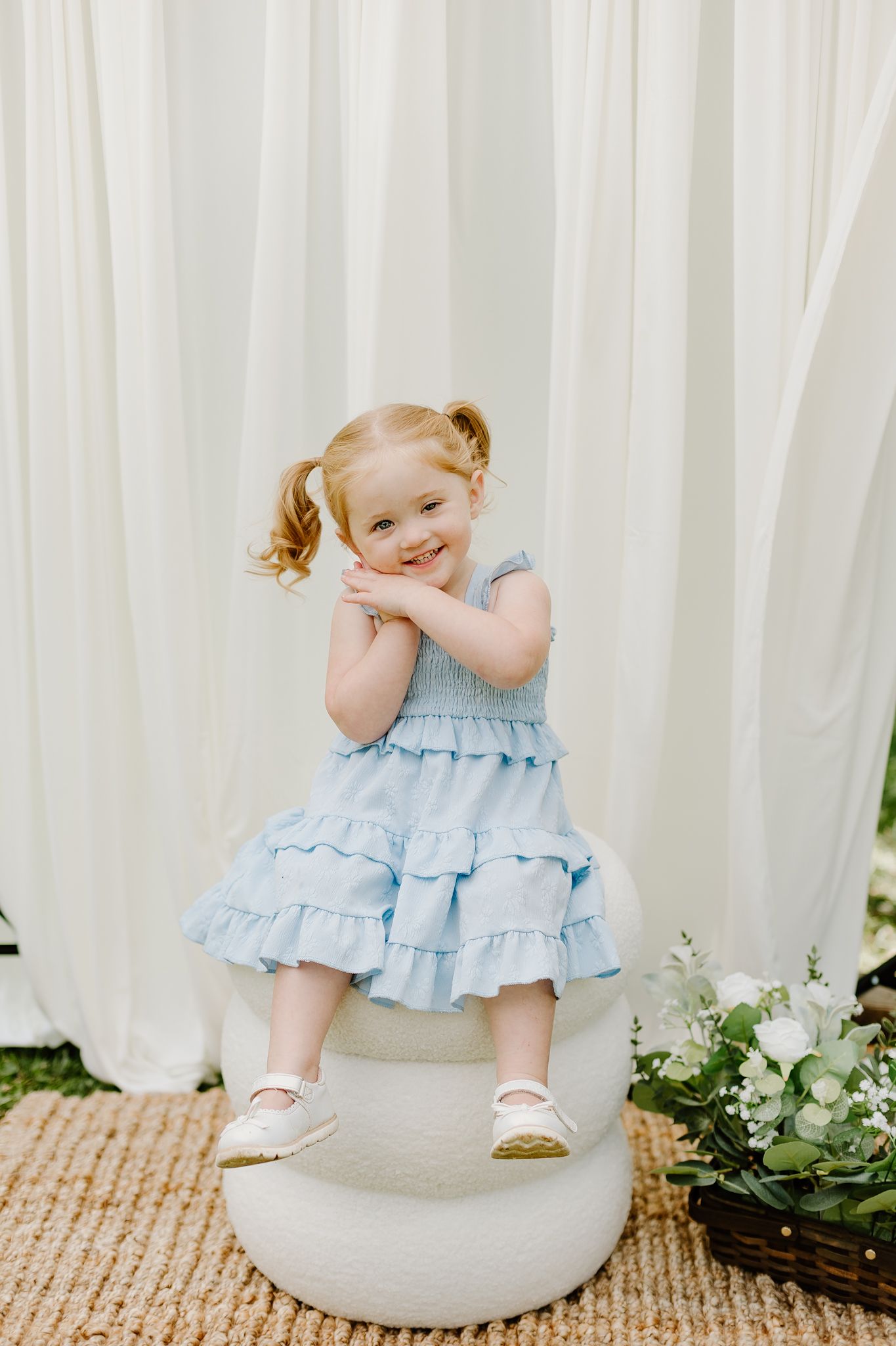 Smiling toddler girl in a light blue ruffled dress with pigtails, standing on a white ottoman outdoors.