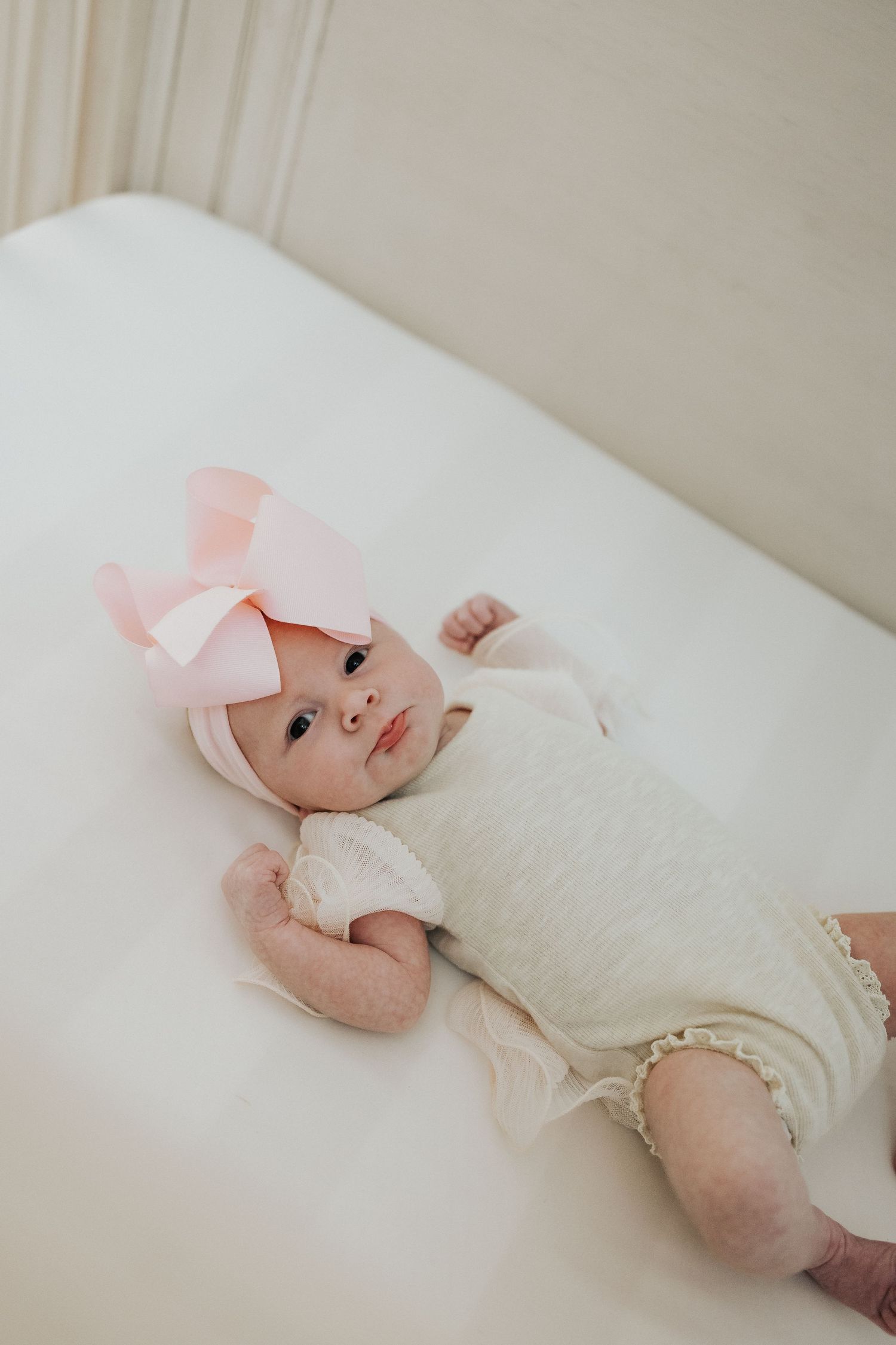 Adorable baby girl lying on a white surface wearing a pink bow headband.