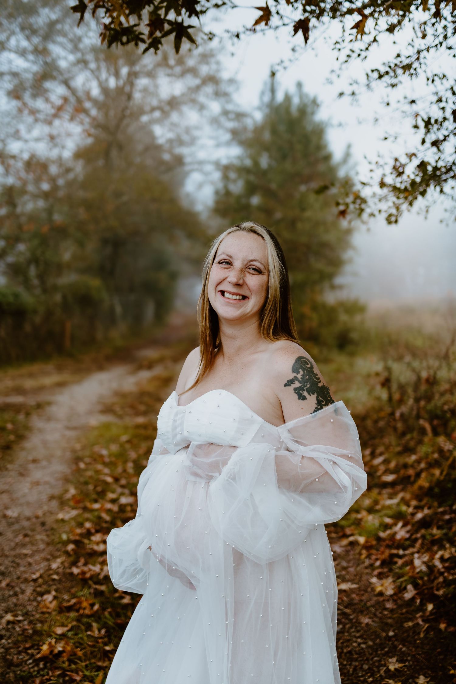 Laughing pregnant woman in white sheer gown on misty autumn woodland path.