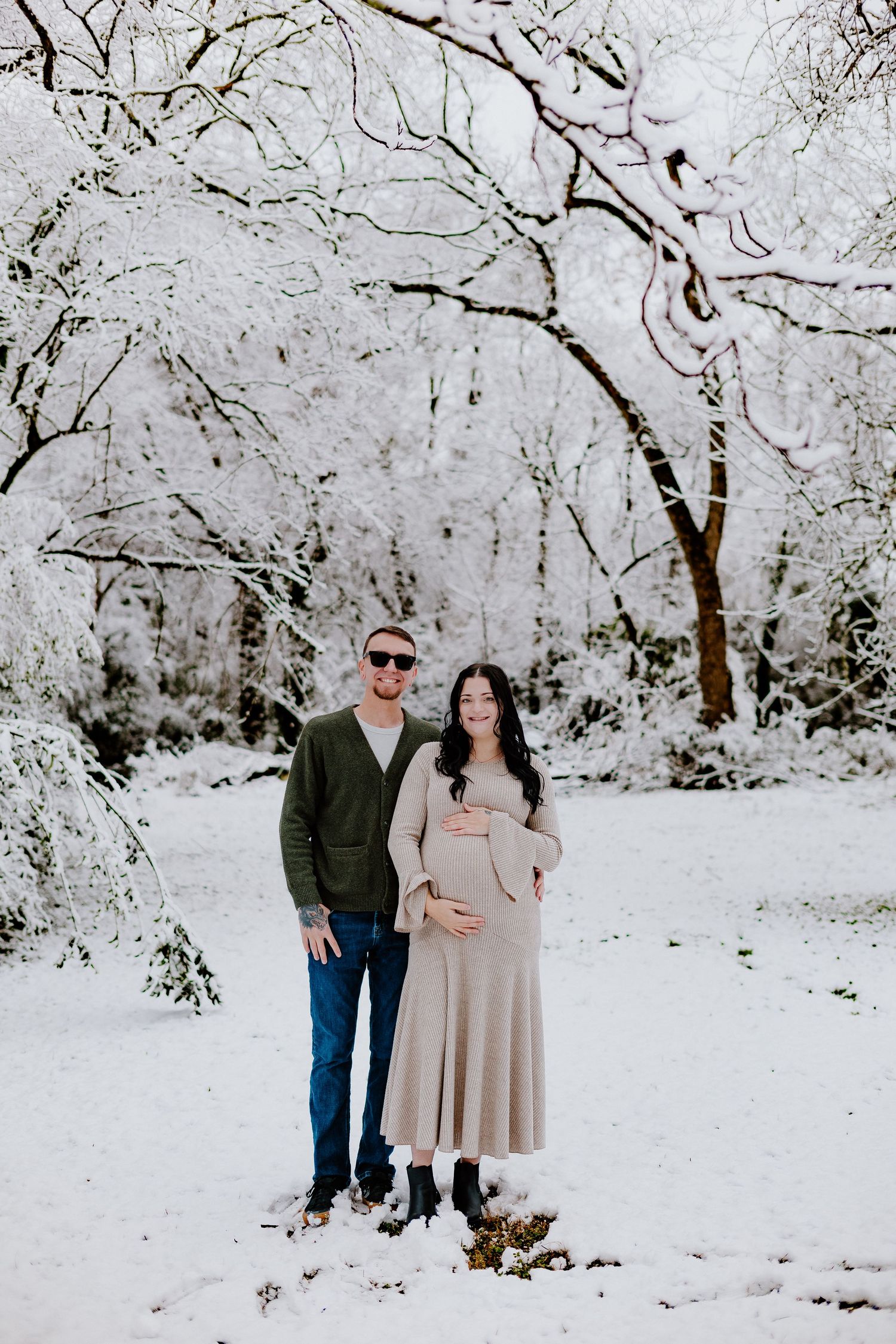 Couple poses in snowy winter wonderland, woman in beige maternity dress cradling baby bump.