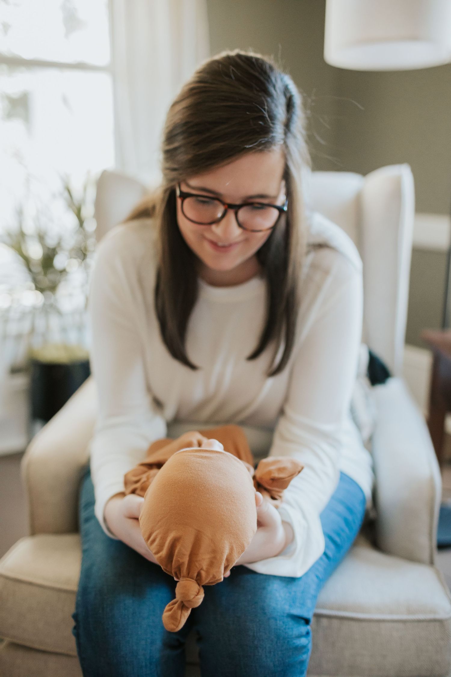 Smiling mother with glasses sits in armchair lovingly cradling her newborn baby in brown outfit.