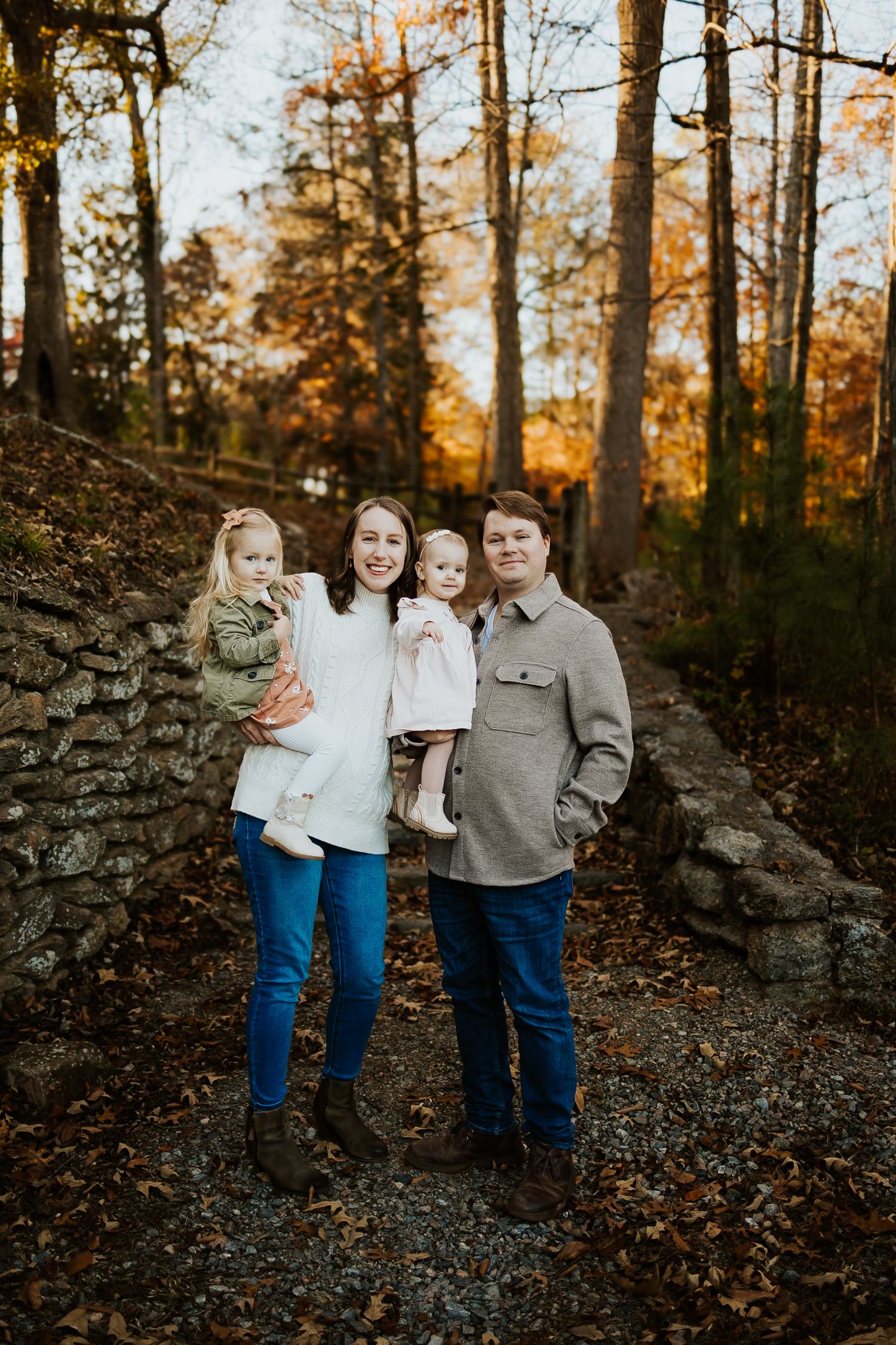 Young couple holding two toddler girls in a forest with vibrant autumn leaves at golden hour.