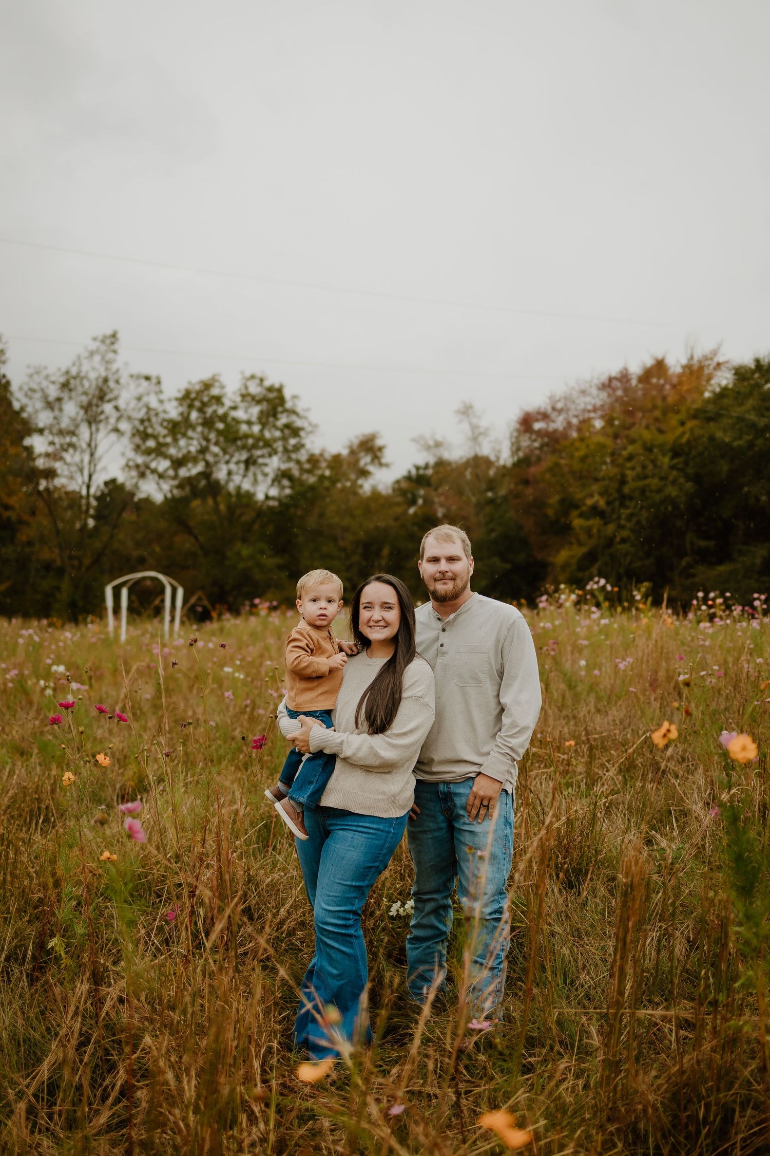 Young family of three poses in a colorful wildflower meadow on an overcast autumn day.