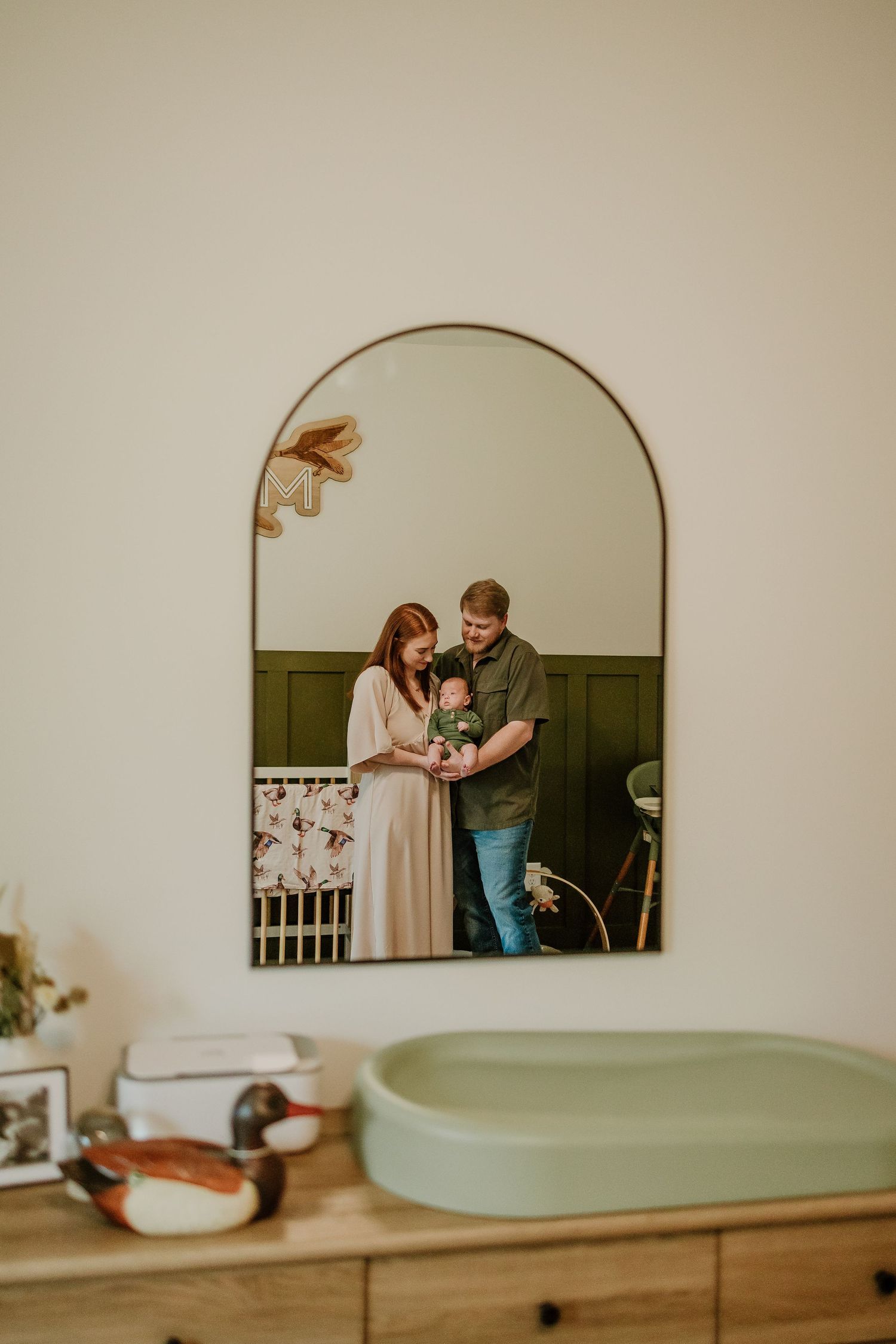 Arched mirror reflection of new parents admiring their newborn baby in a green nursery.