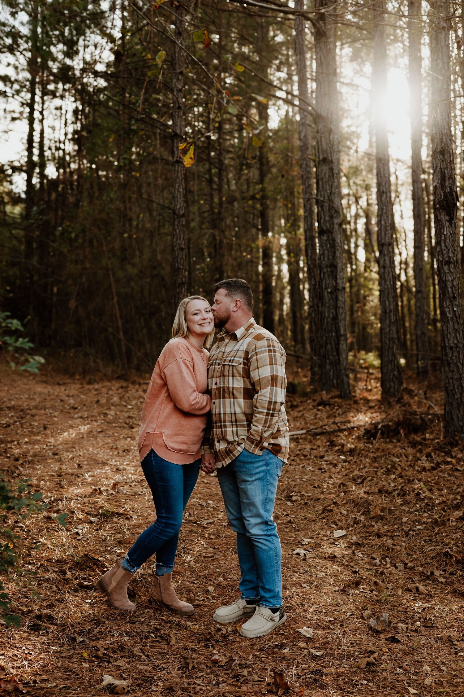 Romantic couple sharing a tender kiss on a sunlit forest trail surrounded by tall pine trees.
