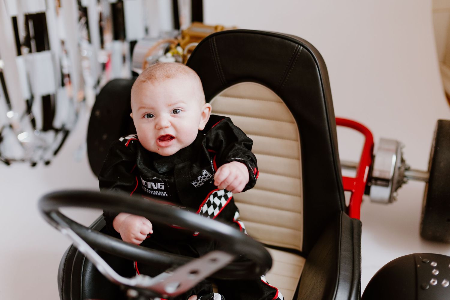 Close-up of smiling baby in black racing suit seated behind steering wheel of red go-kart.