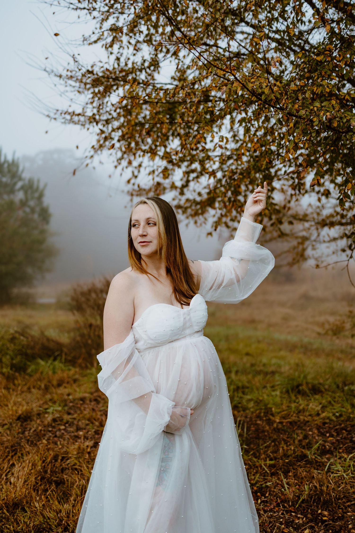 Pregnant woman in sheer white gown stands beneath autumn tree in misty outdoor setting.