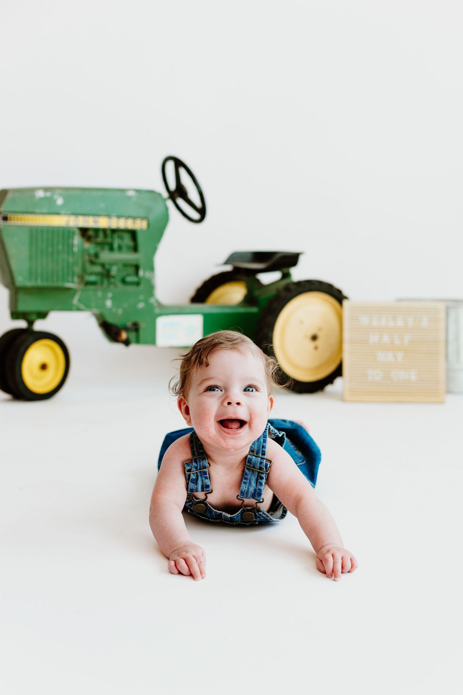 Happy baby in denim overalls doing tummy time near John Deere tractor at half birthday session.