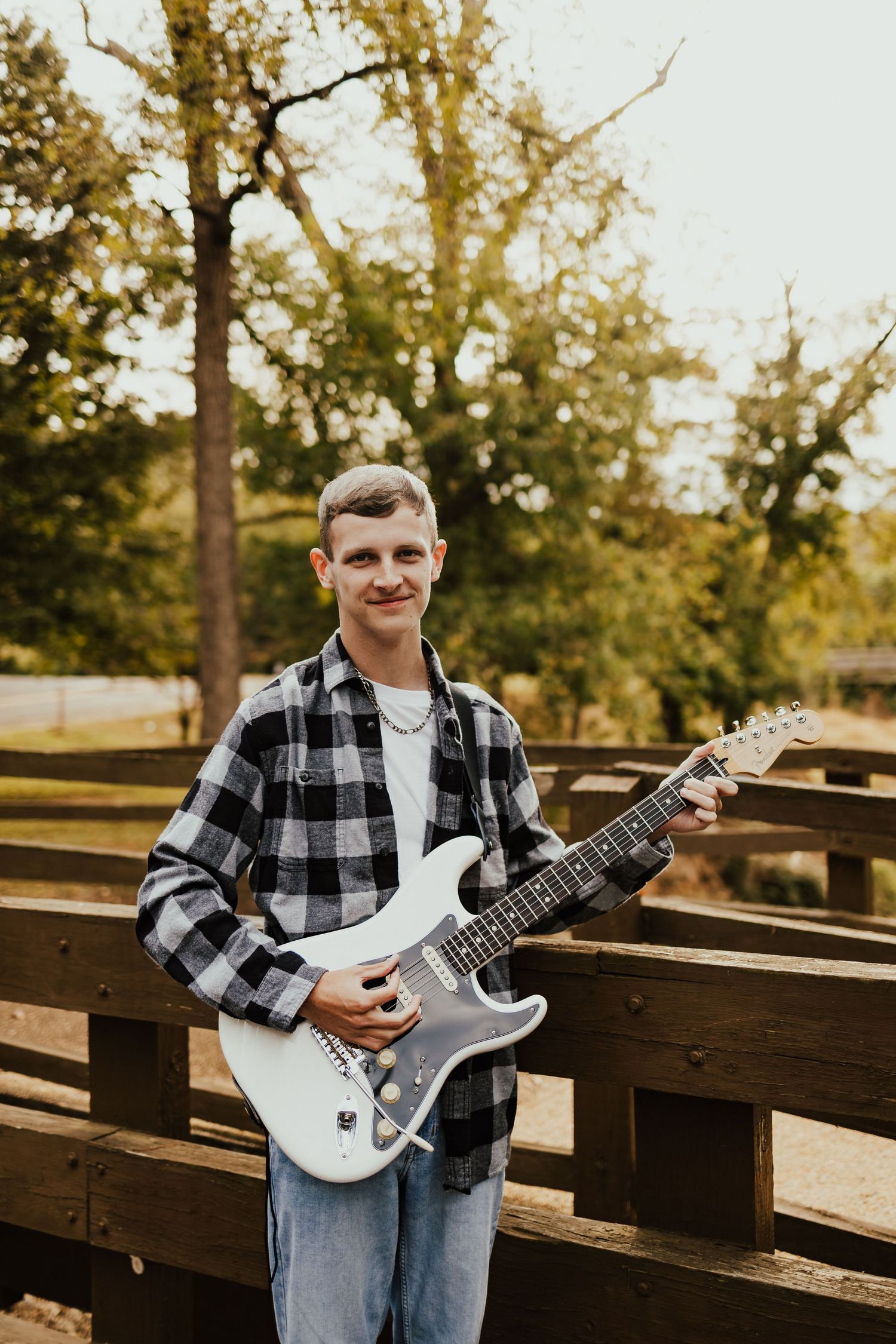 Teenage boy holding white electric guitar wearing plaid shirt, standing on wooden bridge outdoors.