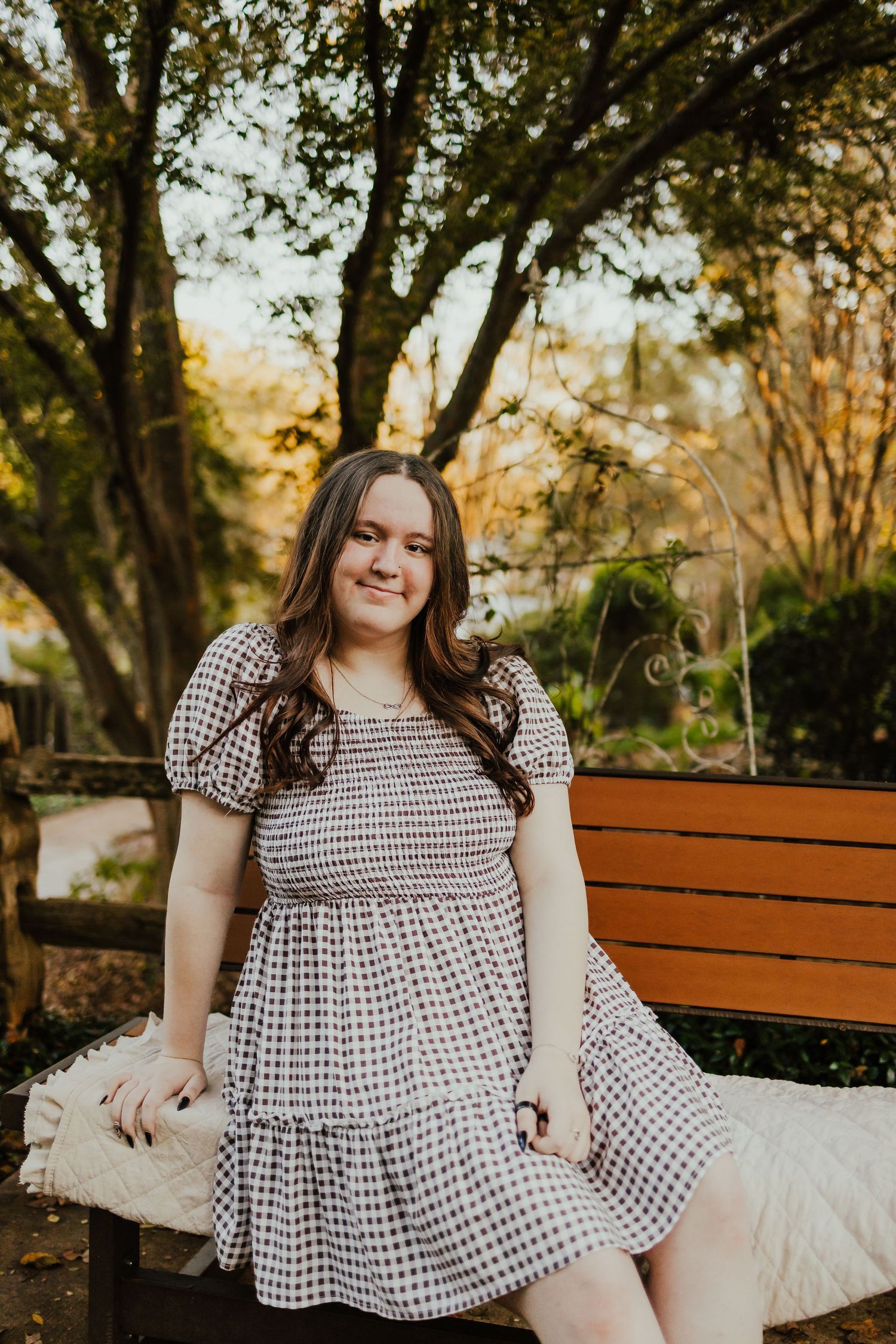 Young woman in gingham dress sitting on wooden bench surrounded by autumn trees at golden hour.